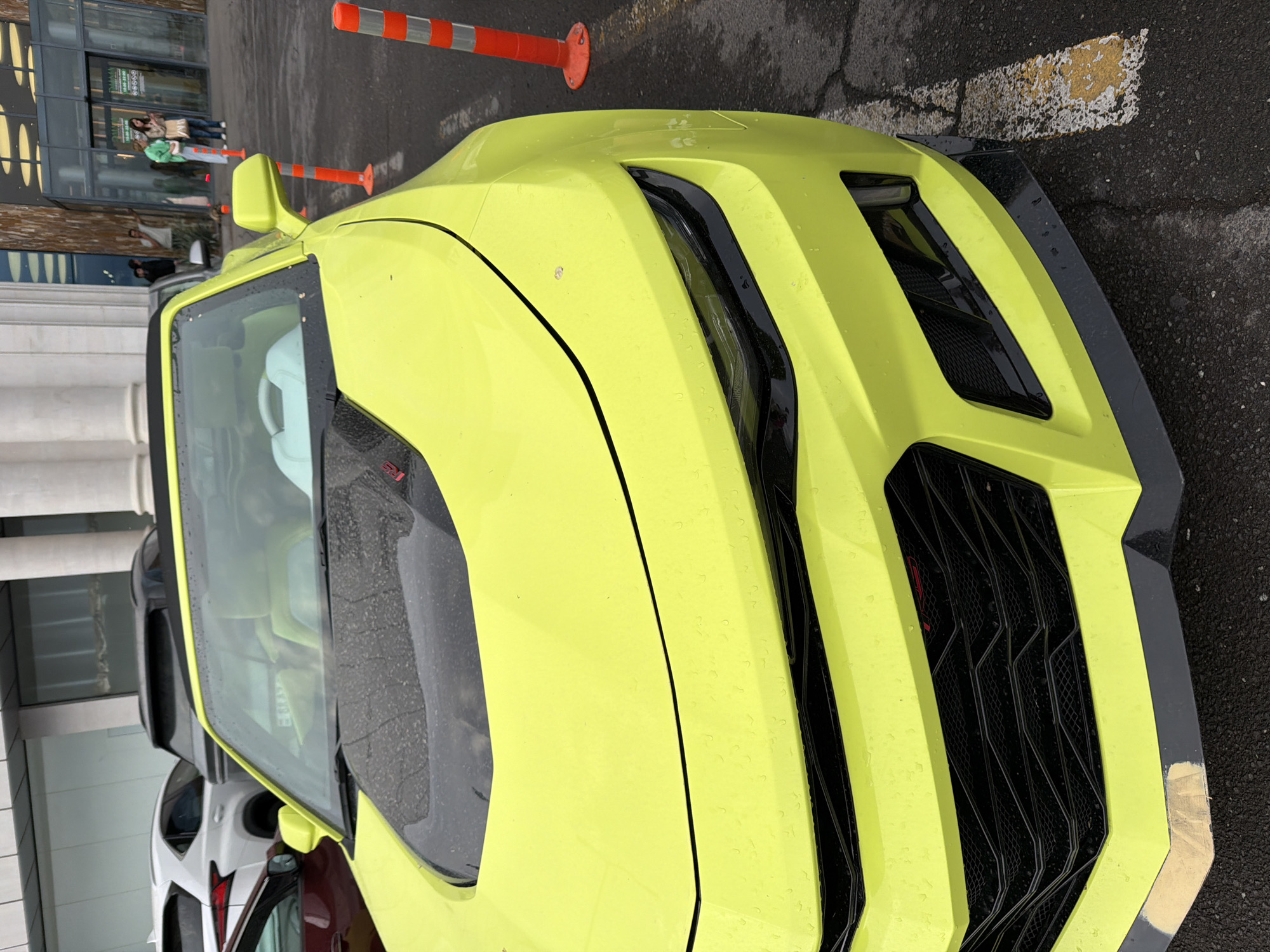 The image captures the front of a bright yellow sports car parked on an asphalt surface, possibly in Yerevan, Armenia, based on visual markers. The car's design is aggressive, with sharp angles, a prominent black grille, and distinctive headlights. The car appears to be wet, possibly from rain, with water droplets visible on its surface.
In the background, blurred figures suggest pedestrians near a building, and orange and white traffic cones are visible. Other vehicles are partially seen to the left of the yellow car. The overall environment seems urban and somewhat overcast.