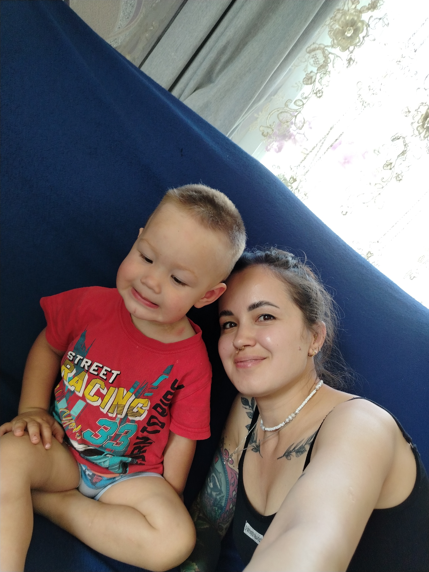 In Гожан, Russia, a young child and a woman are seated on a blue sofa. The child, wearing a red t-shirt with a "Street Racing 33" graphic, is positioned to the left. The woman, on the right, is wearing a black tank top and a beaded necklace, with visible tattoos on her arm. A sheer curtain with floral embroidery is visible in the background, letting in natural light.