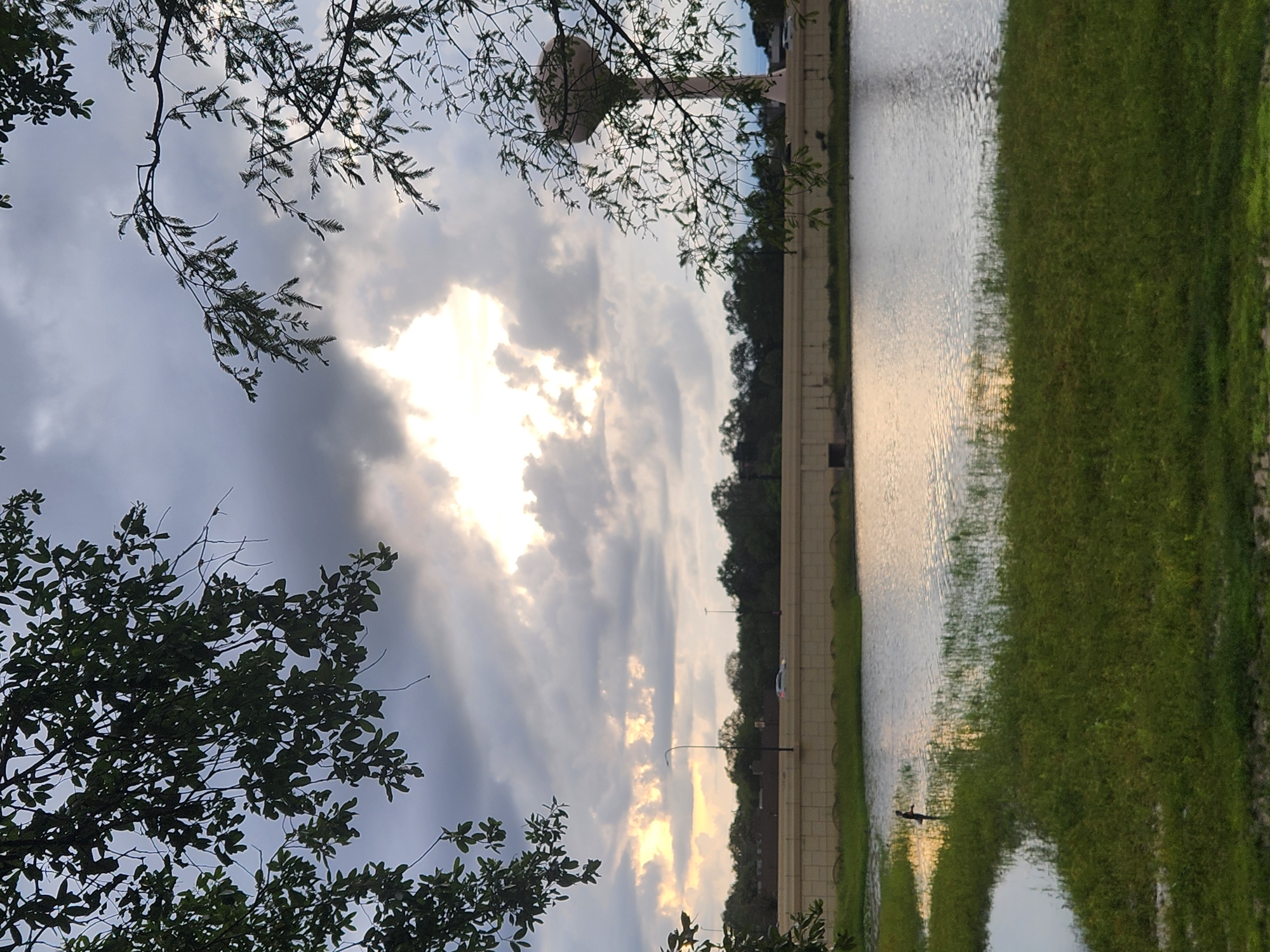 This image depicts a view of the Altamonte Springs water tower from across a body of water. The scene is characterized by a cloudy sky with the sun breaking through in the center, illuminating the clouds. In the foreground, there is a grassy bank leading to a calm body of water. A small figure, possibly a person, stands in the shallow water near the bank. Across the water, a concrete barrier and a road are visible, with a few cars passing by and streetlights. The Altamonte Springs water tower stands prominently in the background to the right, silhouetted against the bright sky. Trees line the horizon behind the road. The image appears to be taken during late afternoon or early evening, given the lighting conditions and the colors in the sky.