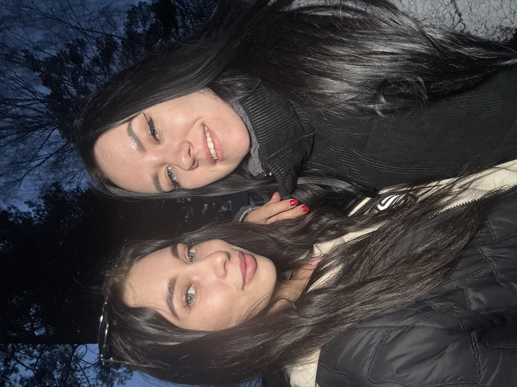 Two women are posing for a selfie outdoors at night. The woman on the top is smiling, wearing a black sweater with a textured knit, and has long, dark hair. The woman on the bottom is also smiling and has long dark hair. She is wearing a quilted jacket with white trim and sunglasses are visible next to her face. Behind them, silhouetted tree branches can be seen against a dark blue sky. There is no information about location of the scene.