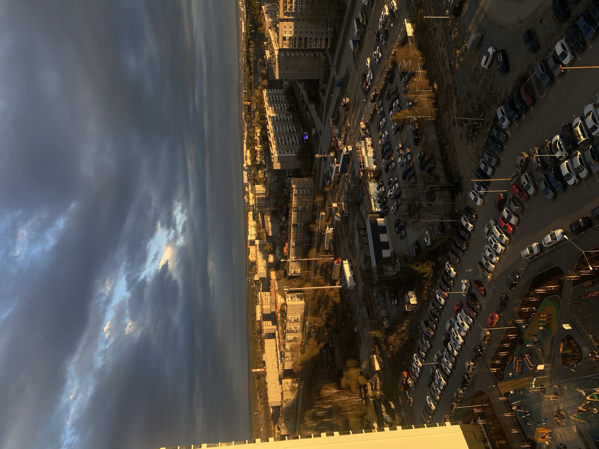 The image, taken from a high vantage point, presents an aerial view of a dense urban landscape, likely Yekaterinburg, Russia, under dramatic sky conditions. The scene is illuminated by the warm, low light of either sunrise or sunset, casting long shadows across the environment.
The sky, occupying the upper half of the correctly oriented image, is predominantly covered by heavy, dark grey clouds, hinting at recent rain or an approaching storm. However, patches of lighter, brighter clouds and glimpses of blue sky break through, catching the golden light, especially towards the horizon.
Below the sky lies the city. Numerous multi-story residential buildings, characteristic of Soviet-era architecture, are visible, densely packed together. Many of these buildings are lit from the side by the low sun, highlighting their facades in warm tones, while other sides remain in shadow. The landscape is interspersed with a grid of roads and extensive parking lots, which are filled with a multitude of cars, mostly sedans and hatchbacks of various colors. The parked vehicles are also struck by the golden light, creating reflections and distinct shadows.
Sparse vegetation, including trees with bare branches, suggests the time of year is late autumn or early spring. In one area, a colorful children's playground with slides and other structures is visible, nestled among the buildings and parking spaces. The playground appears empty.
The overall impression is one of an ordinary urban scene captured at a magical hour, where the interplay of dark, stormy clouds and golden sunlight creates a striking contrast and a sense of calm. No visible text is present in the image.