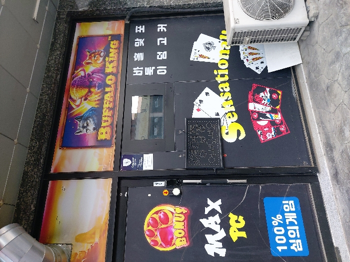 The image depicts the exterior of a commercial storefront in Gimpo-si, South Korea. The dark-colored facade is adorned with various graphics and text, indicating an entertainment or gaming establishment.
At the top, a large banner displays "BUFFALO KING" in gold lettering, positioned below vivid images of a gray wolf, a yellow buffalo, and a brown cougar against a stylized purple and orange sky. To the immediate left of this banner, desert-like artwork extends across the upper section of the storefront. A silver metal ventilation pipe is prominently visible in the upper left corner.
The two main doors below are dark. The left door features "BONUS" in red and yellow lettering, a large yellow paw print graphic, and "MAX PC" in yellow. A blue rectangular sign on this door reads "100% 심의게임" (100% recreational game) in white Korean text.
The right door or window section showcases "Sensational" in yellow script, along with several graphics of playing cards, including various poker hands and aces of spades. A small, black LED sign, illuminated with white dots, displays "영업중" (Open for Business) in Korean. Additional Korean text, "바둑 맞포 이 엄 고커," is present above this section. A small security emblem is affixed near the top center of the door frame.
An external white air conditioning condenser unit is positioned on the concrete pavement to the far right of the scene. The ground is a textured concrete surface. No individuals are present in the image.