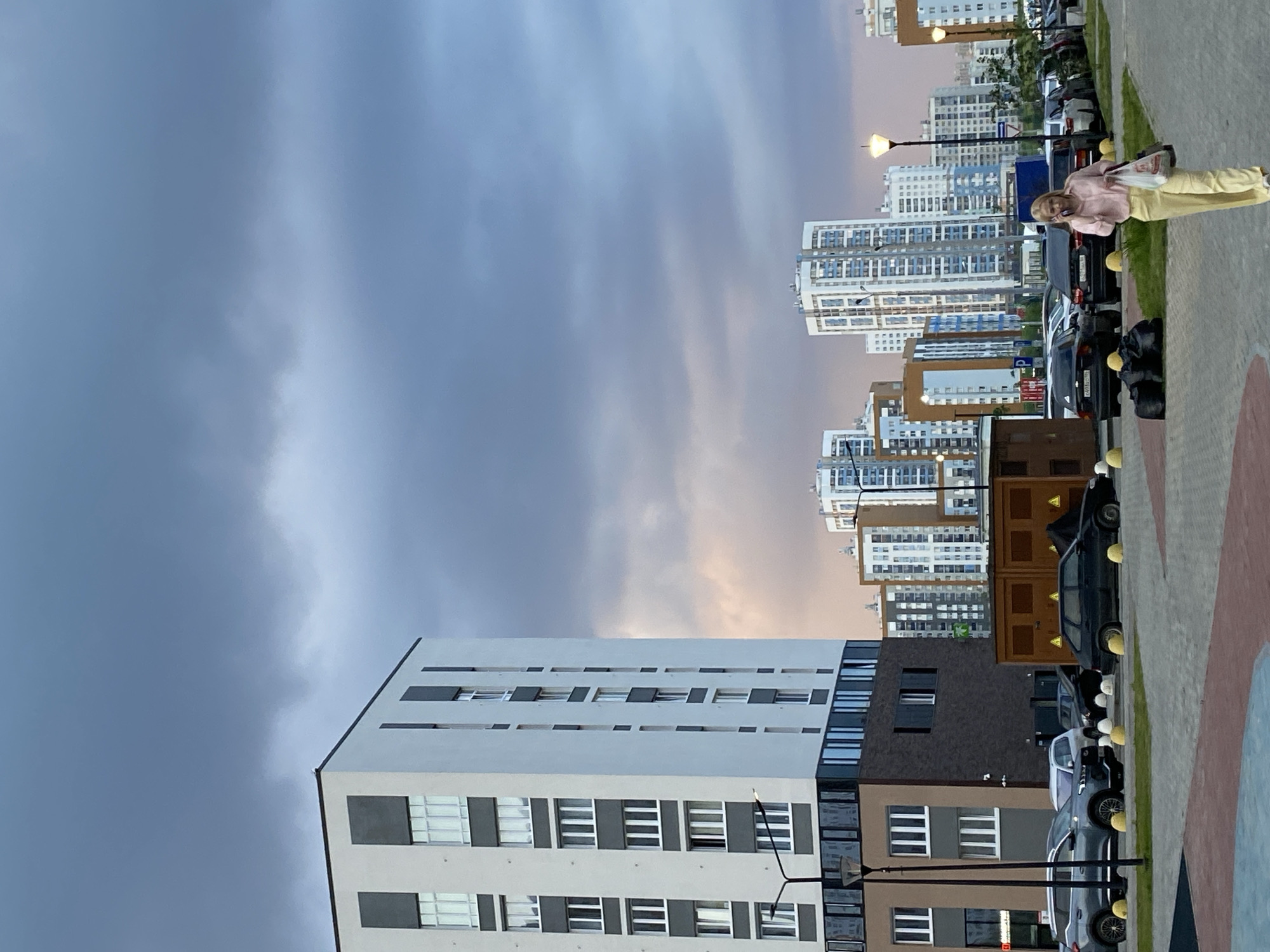 The image shows a street scene in Yekaterinburg, Russia, with a line of modern, multi-story residential buildings in the background. In the foreground, a woman with blonde hair, wearing a pink blouse and yellow pants, is walking and speaking on her cell phone. She is carrying a plastic bag in her right hand. Several parked cars are visible, including a black sedan and a grey SUV. The sky is overcast with dark clouds, suggesting impending rain or an evening setting. The architecture of the buildings is contemporary, featuring large windows and geometric designs. A street lamp stands near the left side of the image.
