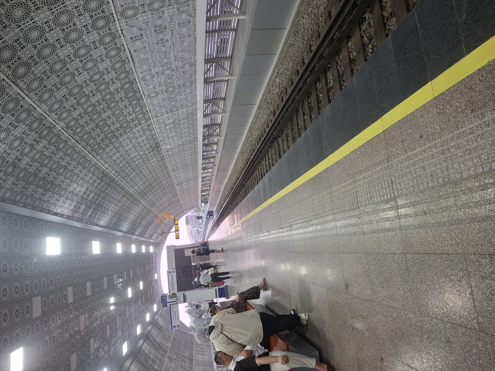 This is a photograph taken at a metro station in Tashkent, Uzbekistan. The station appears to be underground, with an arched ceiling decorated with intricate patterns. The platform is made of granite tiles and has a yellow safety line bordering the tracks. People are waiting on the platform, some sitting on benches and others standing. The train tracks are visible to the right of the platform, with gravel ballast and a metal fence in the background. A digital display board shows the time and possibly train information. The overall impression is of a modern and clean public transportation facility.