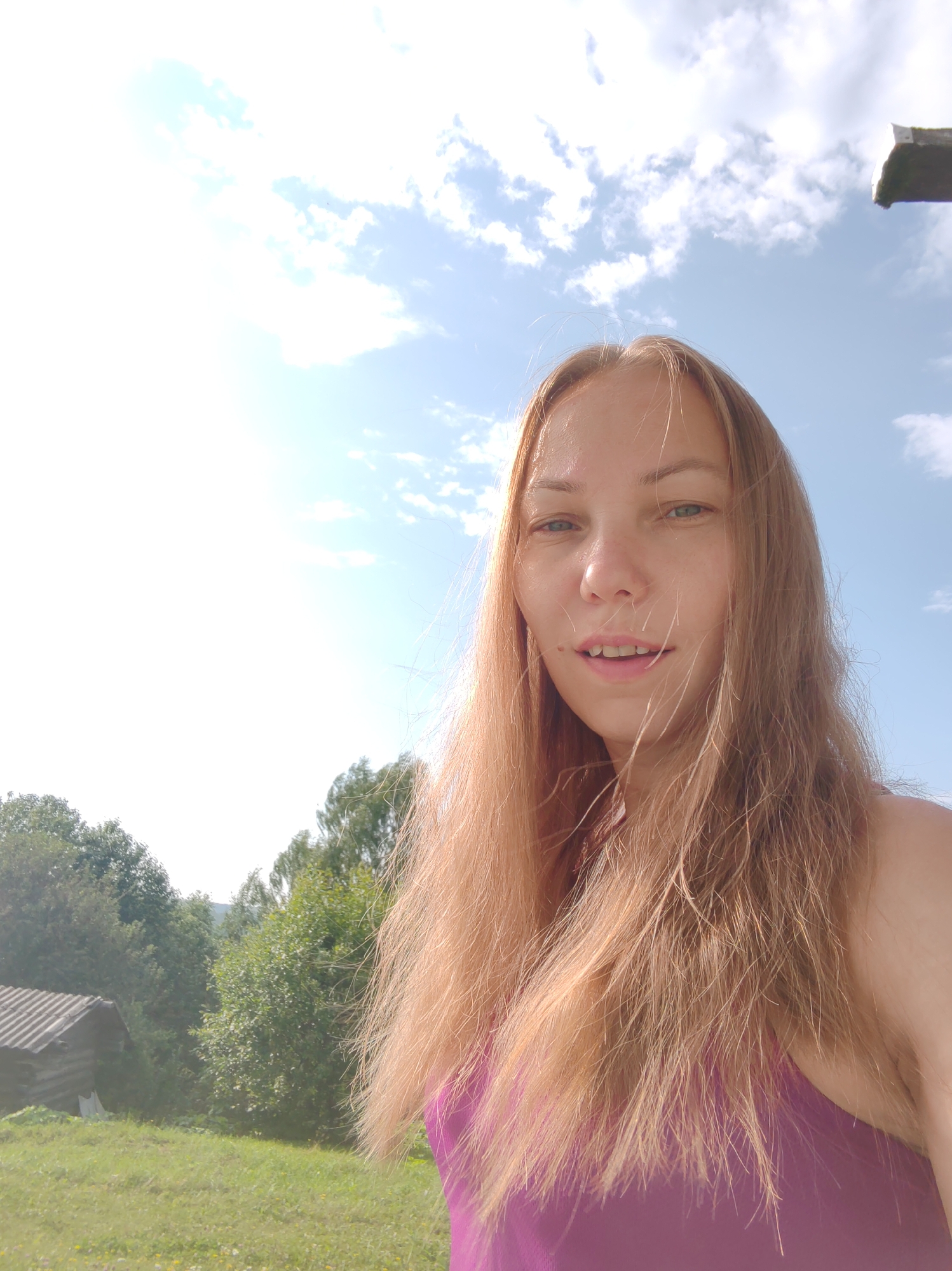 A young Caucasian woman with long, light brown hair, wearing a purple tank top, takes a selfie outdoors. She has fair skin and green eyes, with her mouth slightly open. The background suggests a rural setting in Чухлэм, Russia, featuring a grassy field, lush green trees, and a rustic wooden building with a gray roof. The sky is bright blue with scattered white clouds, and the sun is shining, casting a bright glare on the left side of the image. A dark, wooden structure is visible at the top right corner.