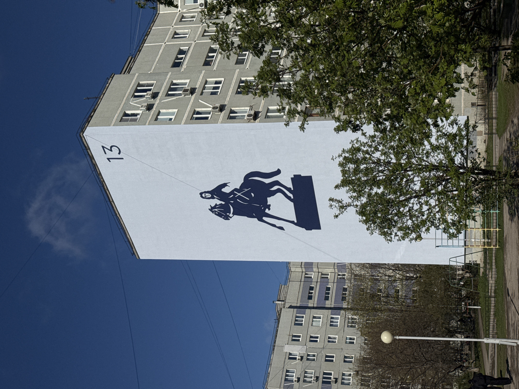 The image captures a tall, light-grey residential building under a bright blue sky, adorned with a large mural or banner on one of its end walls. The mural, colored light blue or light grey, features a prominent dark silhouette of a person on horseback, mid-jump over a rectangular obstacle or pedestal. The number "13" is clearly visible in the upper left corner of this mural.
The building itself is multi-storied with numerous rectangular windows, some of which have external air conditioning units. Lush green trees with full foliage are scattered across the scene, with some branches extending into the frame, partially obscuring the building and the mural.
In the background, on the right side, another similar-looking grey residential building is visible, also partially hidden by trees. The ground level shows a paved path or dirt area, a metal fence, and a yellow and blue playground structure. A white lamppost with a spherical light fixture stands in the lower right, and a dark, indistinct figure, possibly a person, can be seen walking near its base.
The scene appears to be set on a clear, sunny day, likely during spring or summer, given the vibrant green leaves on the trees. The overall impression is that of an urban or suburban residential area.