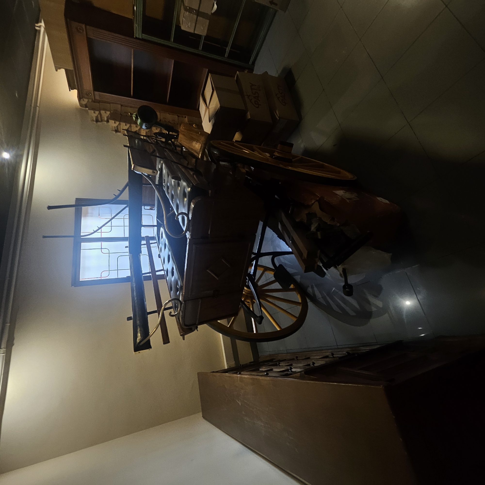 The image depicts the interior of what appears to be a museum or exhibition space in Almadrones, Spain. The central focus is an antique horse-drawn carriage, likely a vis-à-vis or similar design, characterized by its wooden body, leather seating, and large spoked wheels. The carriage is positioned in a room with light-colored walls, illuminated by artificial lighting that creates shadows on the tiled floor. To the left of the carriage, a dark wooden cabinet or dresser with multiple drawers is visible. In the background, a large window with a grid pattern is present, allowing some diffused light to enter. Further to the right, a tall, dark wooden bookshelf with several shelves is seen, and stacks of cardboard boxes, some labeled with "PASTY," are placed nearby. The overall environment suggests a preserved historical setting, possibly showcasing transportation history or local heritage within Almadrones. No people are present in the scene.