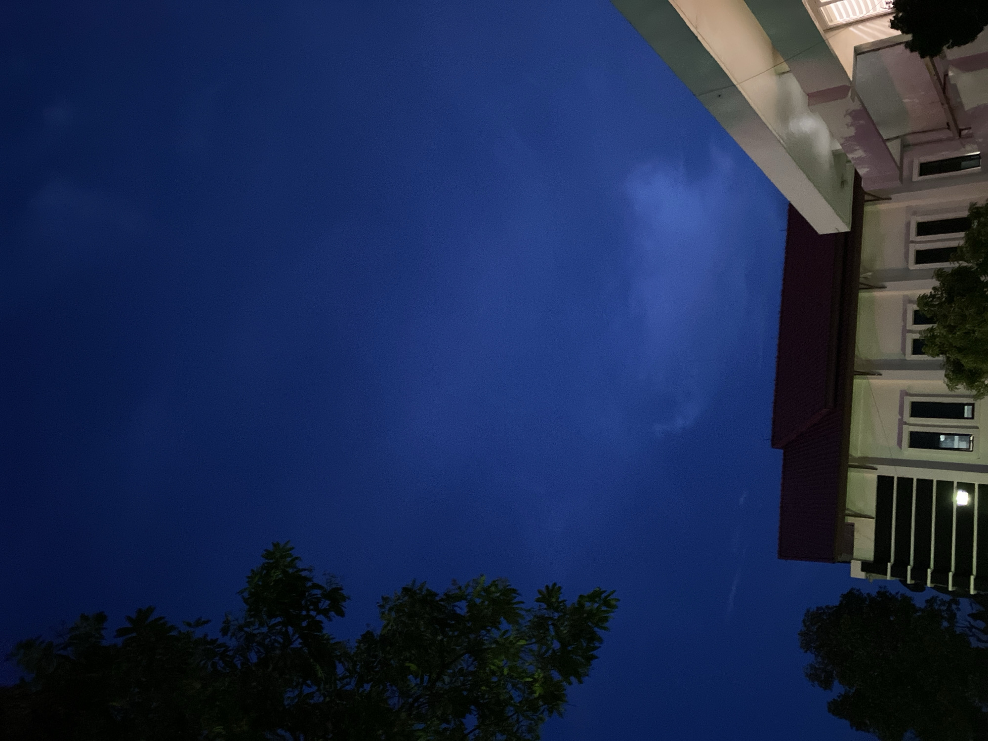 The image depicts a building with a red roof and white walls, situated in a dark blue sky. A tree with dark green leaves is visible in the foreground on the left side, its branches reaching towards the sky. A portion of another building with a white awning and light purple accents is present on the right side of the image. The sky is clear with a few wispy clouds. The overall scene is calm and serene, with the lighting suggesting it is dusk or early evening. No people or specific actions are discernible. The location is identified as Unknown, Unknown.