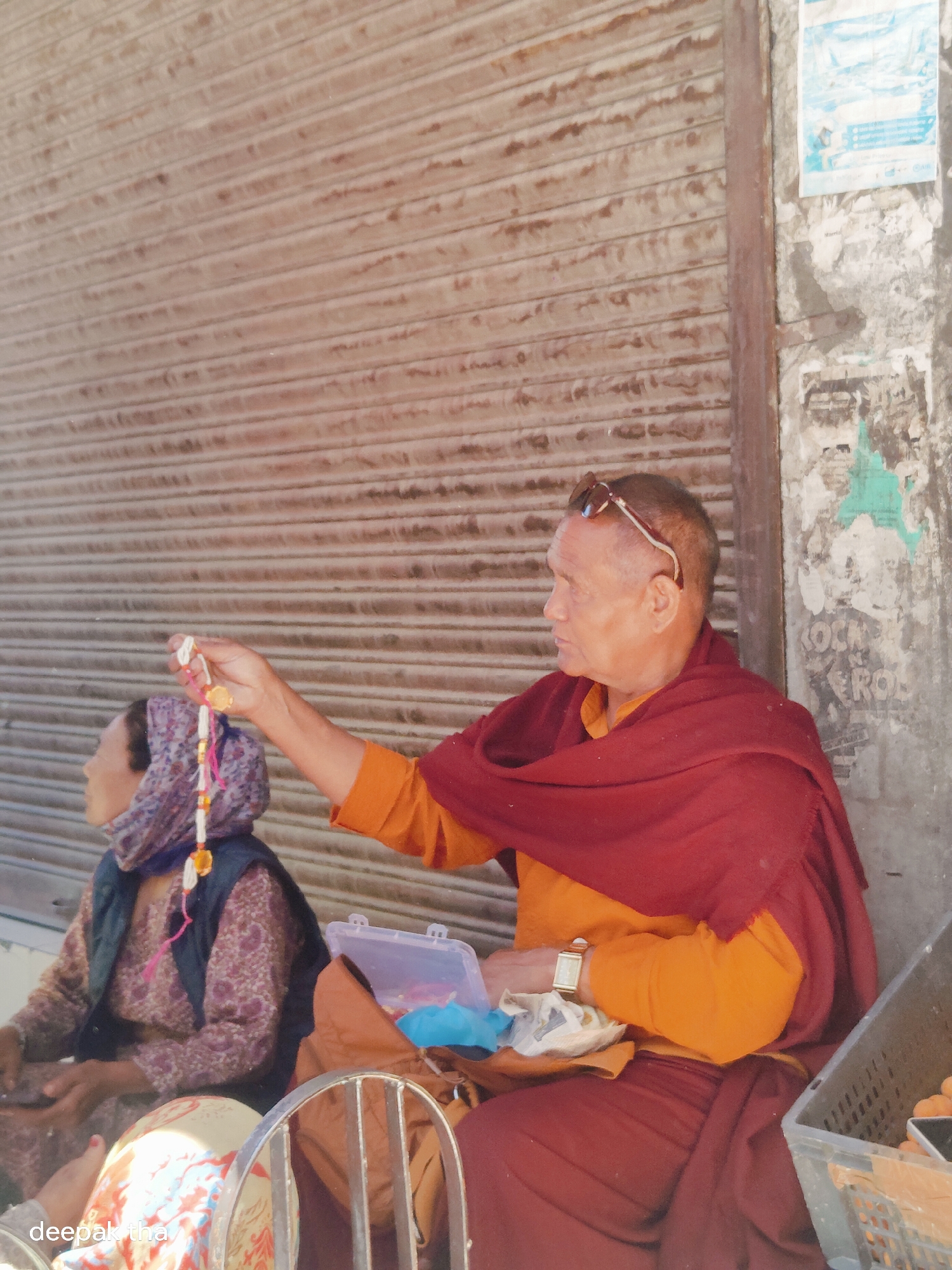 The image captures a scene in the city of Leh, India, with a focus on two individuals. In the foreground, a man, appearing to be a Buddhist monk, is seated and wearing traditional orange robes with a deep red shawl draped over his shoulders. He has sunglasses perched on his head and is holding up a string of prayer beads. To his left, a woman is also seated, wearing a patterned headscarf and a dark vest over a floral dress. The background features a weathered, corrugated metal shutter, characteristic of many shops in Leh, and a distressed wall with peeling posters. A plastic container with some items inside is visible next to the monk, and a crate with what appear to be orange fruits is to his right, suggesting a marketplace or street vendor setting. The lighting indicates it is daytime, possibly late afternoon, with shadows cast by the subjects.