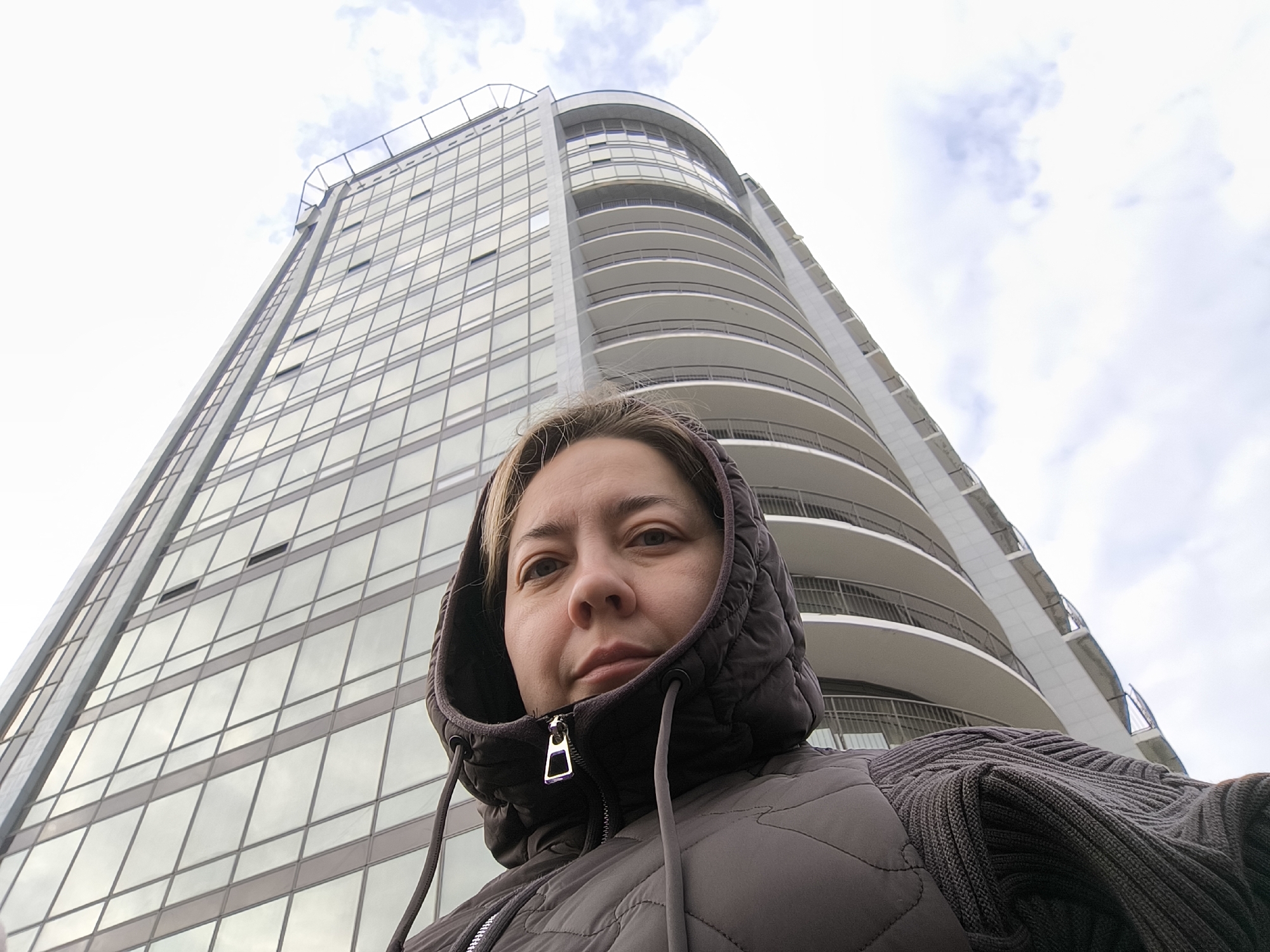 An adult female is depicted in the foreground, facing the camera directly. She wears a dark, hooded, quilted vest over a dark ribbed sweater; her light-colored hair is partially visible. The background consists of a tall, modern multi-story building in Izhevsk, Russia, captured from a low-angle perspective. The building's left facade is composed of reflective glass panels arranged in a grid, while its right side features several curved, white balconies with dark railings. The sky above is overcast with clouds.