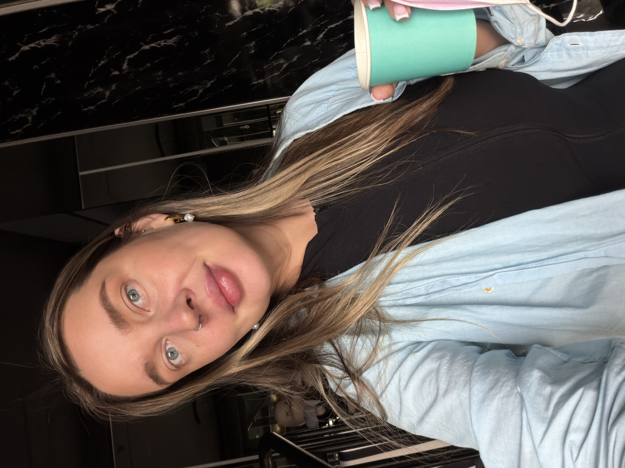 A young woman with blonde hair and blue eyes is holding a blue cup. She's wearing a black top and a light blue shirt. She has a nose piercing and pearl earrings. The background shows what appears to be a black marble countertop and a metal rack. It is not possible to identify the location as Moscow, Russia, from the given image.