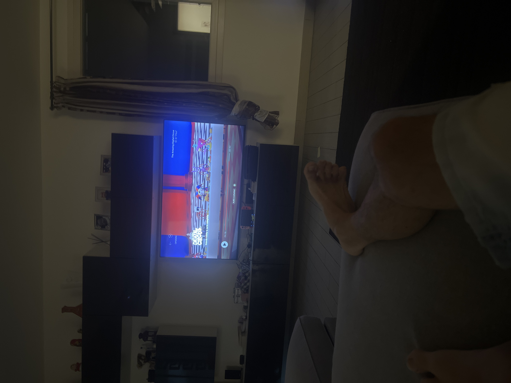 The image captures a living room interior in Arre, Italy, with a focus on a television screen displaying "The Amazing Digital Circus." The scene is dimly lit, suggesting it is evening or nighttime. A person's legs and feet are visible in the foreground, positioned on a sofa. The television, mounted on a dark entertainment unit, shows a colorful cartoon with characters. Various decorative items and electronics are placed on shelves and the entertainment unit. A window with a curtain is visible in the background. The floor appears to be made of wood planks.