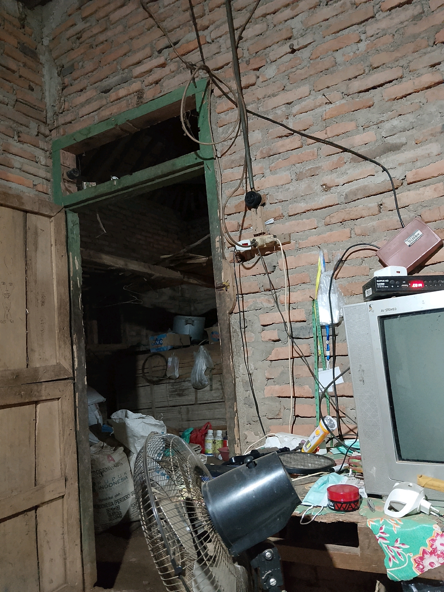 The image depicts a cluttered interior scene, likely a room in a house, with exposed brick walls and a wooden doorframe. The room is dimly lit and appears unkempt. Various objects are scattered around, including a black electric fan in the foreground, a television set, a cable box, what appears to be a router, and several bags of what looks like agricultural supplies or fertilizer. There are also various wires and electrical outlets visible on the wall. The overall impression is one of disorganization and a lack of modern amenities. The specific location of Negeri Agung, Indonesia is not discernible from the visual information presented in the image.