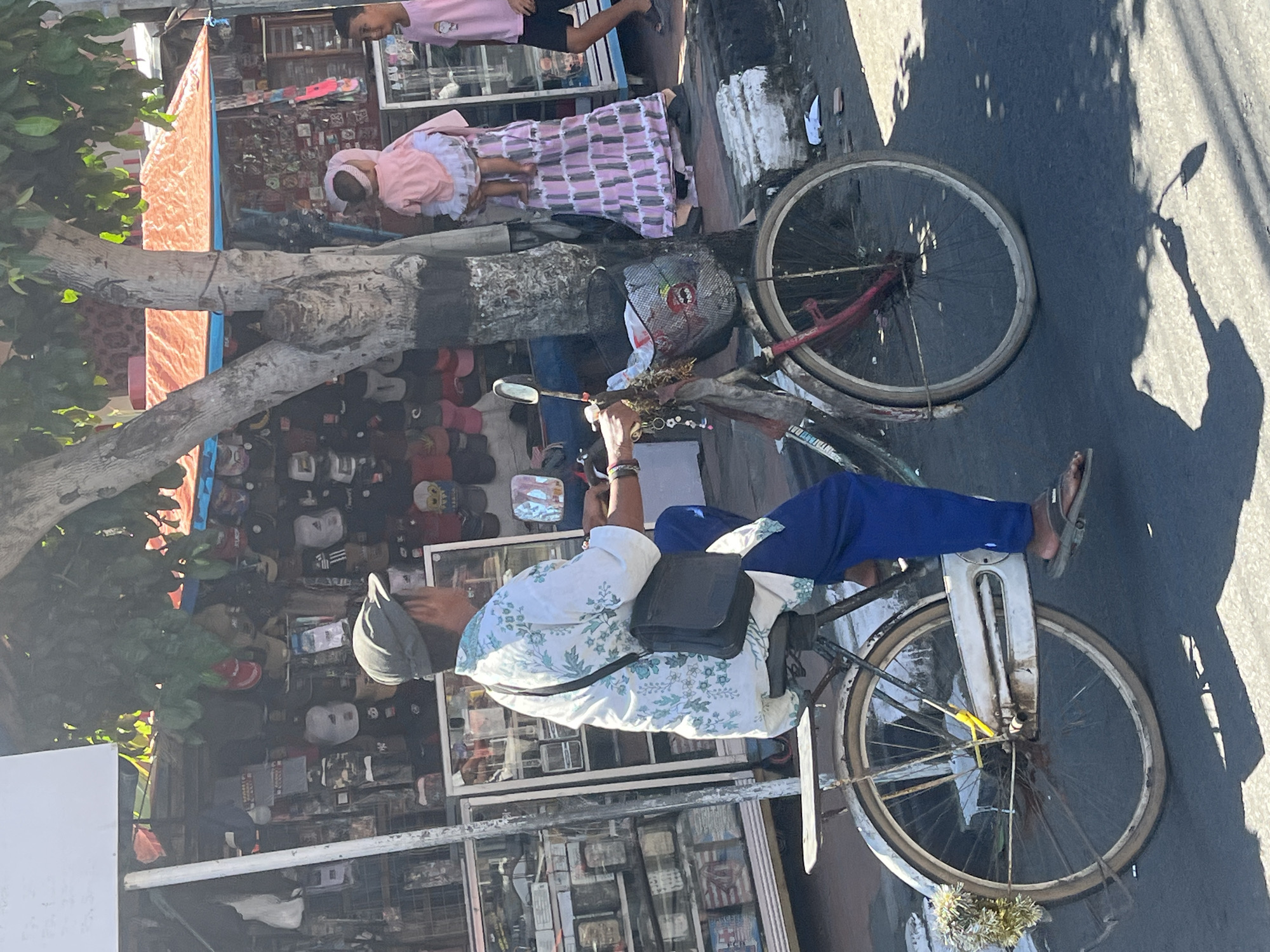 In the image, a man wearing a cap, a floral shirt, and blue pants rides a bicycle on a street in Desa Tambakreja, Indonesia. He has a dark crossbody bag on his shoulder. The bicycle has a basket in the front containing a white bag. To the right of the man, a woman in a pink patterned dress holds a child wearing pink. Further to the right, a young boy in a pink t-shirt walks. Behind them, shops display various items including hats and accessories. The scene is set under a tree with leafy branches, and the ground is paved. The lighting suggests it is daytime, with shadows visible on the road.