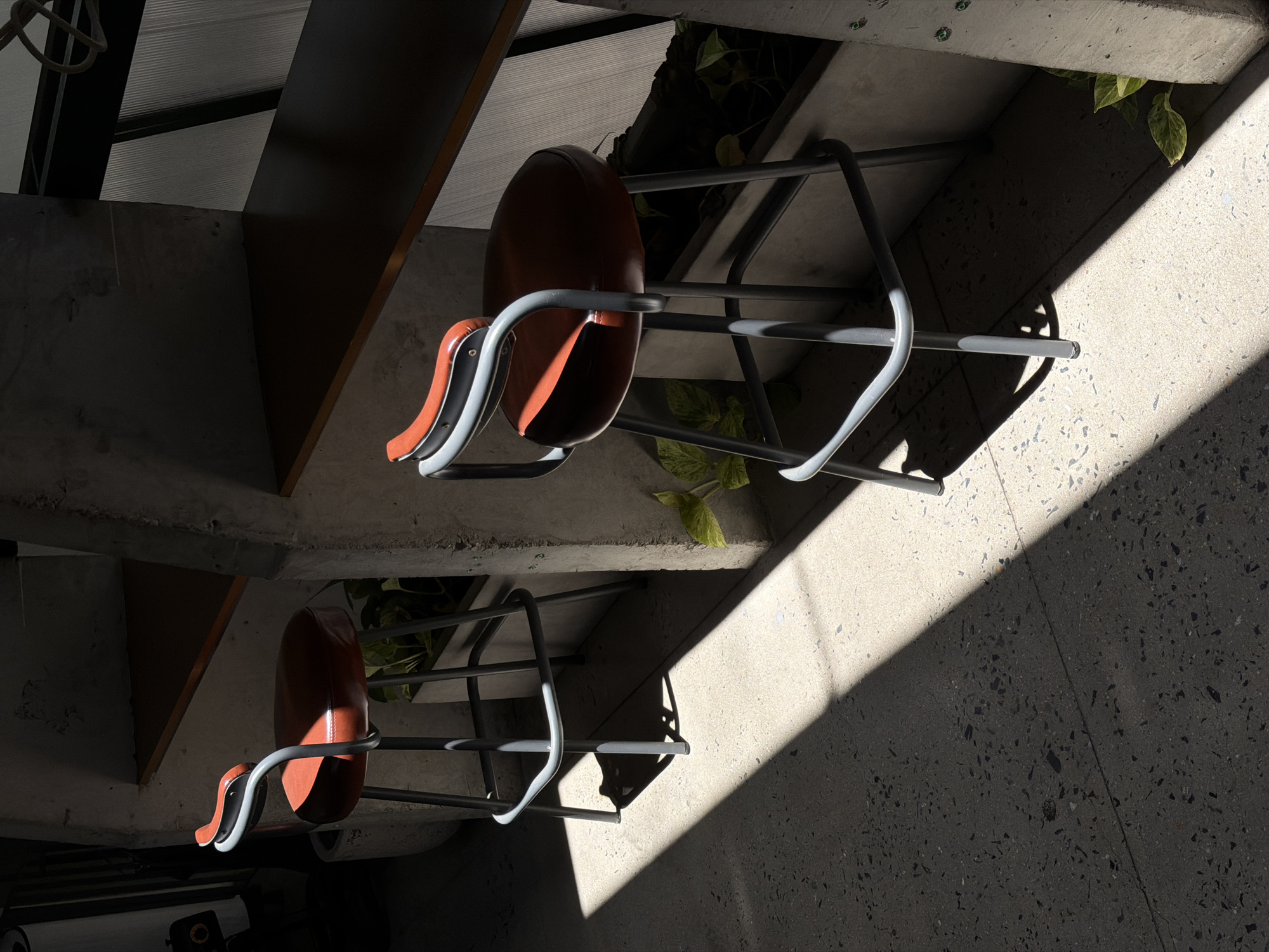 A high-angle shot depicts two brown chairs with gray frames situated beneath a structure, likely stairs. The chairs are identical in design, each having a rounded back and a single armrest. Light is cast diagonally across the scene, creating defined shadows on the floor and highlighting the texture of the concrete surface. Green foliage is visible near the chairs. The scene is likely in Da Nang, Vietnam.