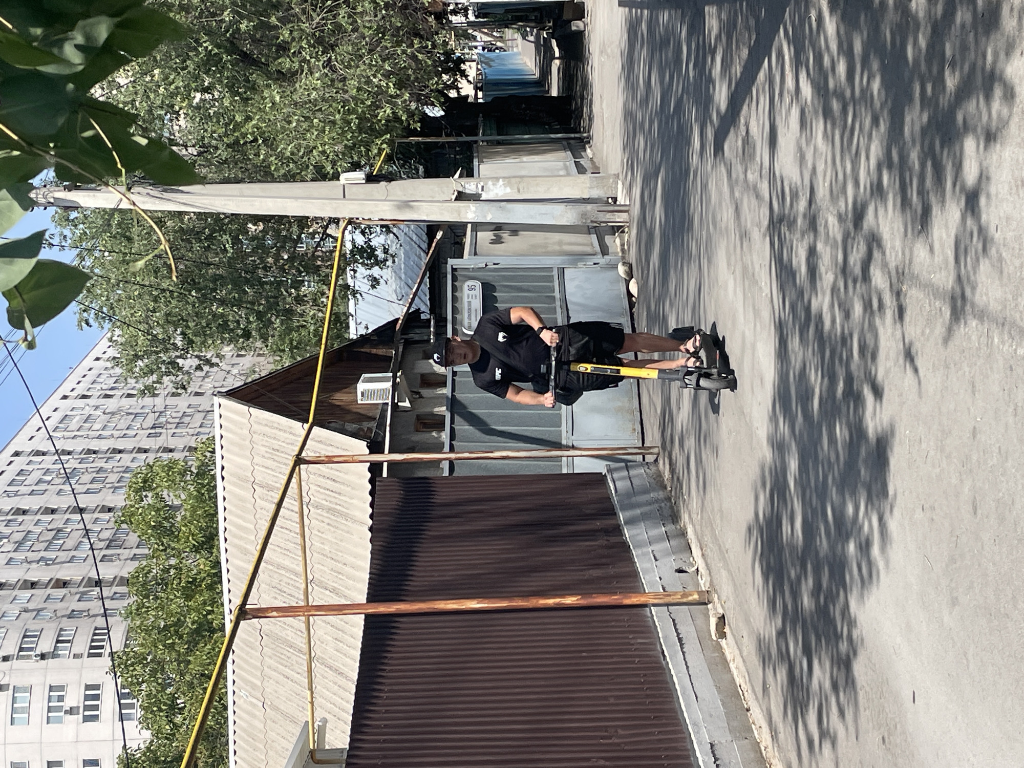 A young Asian male wearing a black t-shirt with a UFC logo, black shorts, a black baseball cap, and sandals rides a yellow electric scooter down a paved alleyway. He is carrying a black bag across his chest. The alley is lined with corrugated brown fences on the left and a mix of grey metal gates and buildings on the right. A large, modern, multi-story residential building is visible in the background, along with trees and a concrete utility pole. The ground is shaded by dappled sunlight filtering through the leaves of trees. The scene appears to be in a residential area in Almaty, Kazakhstan, given the architectural style and the presence of a utility pole with Cyrillic lettering.