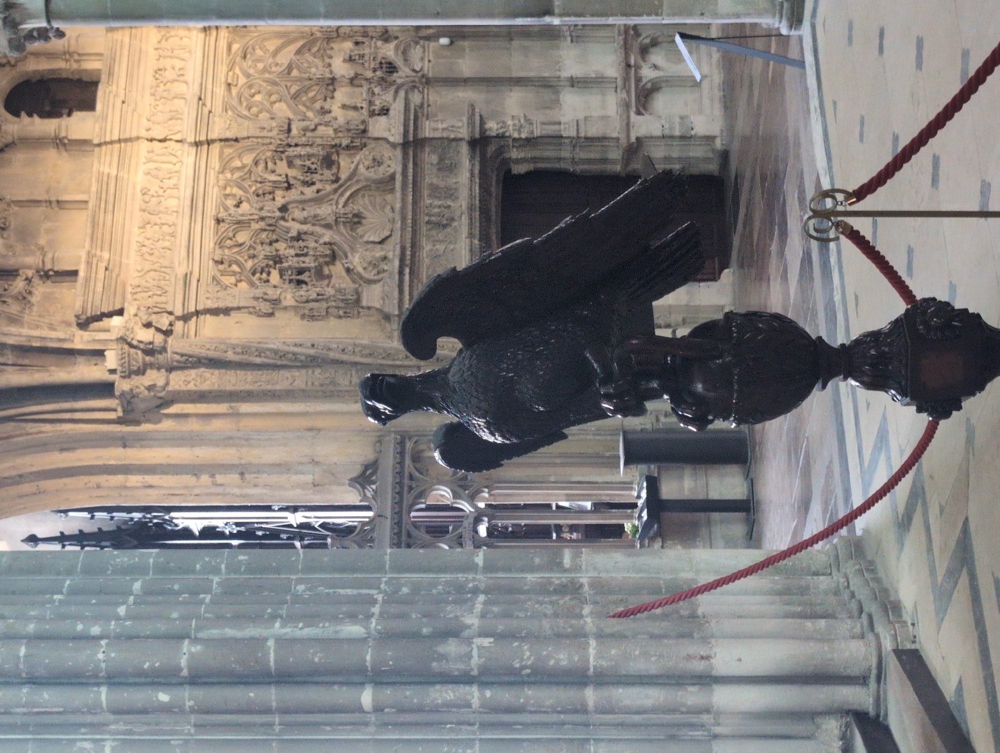 In the interior of a grand stone building, likely a church or cathedral, a dark, ornate eagle sculpture stands prominently in the foreground. The sculpture, with its wings spread, is mounted on a dark, decorative base. In front of the sculpture, a red rope barrier, attached to gold poles, stretches across the scene, delineating a pathway. To the left, a large fluted stone column dominates the left side of the frame. In the background, an elaborate carved stone wall or monument features intricate religious carvings and architectural details. A large arched window or doorway is visible behind the sculpture, with gothic architectural elements. The floor is tiled with a pattern of squares and rectangles. The overall atmosphere is solemn and historical. This scene is located within the church of Saint-Jacques in Le Tréport, France. There are no people visible in the image.