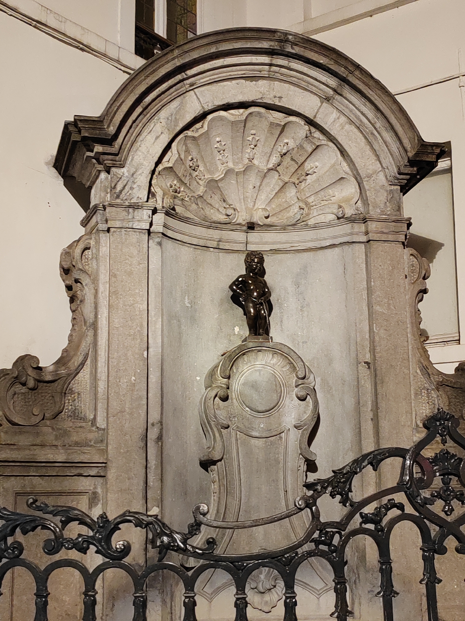 This image captures the iconic Manneken Pis statue in Brussels, Belgium. The small bronze sculpture of a boy urinating stands atop an ornate pedestal, which is itself part of a larger stone fountain structure. The fountain is enclosed by a decorative black wrought-iron fence, adding a touch of historical grandeur to the scene. The statue is centrally placed within a recessed niche featuring a scalloped shell motif at the top, all carved from stone. The overall architecture suggests an outdoor public space, likely a square or a street corner, given the presence of the fence and the implied architectural context. The lighting in the image suggests it might be evening or night, or perhaps an indoor setting with artificial illumination, as shadows are cast distinctly. The statue itself is a well-known landmark and a symbol of Brussels, often adorned with various costumes depending on the occasion. No visible text is present in the image.