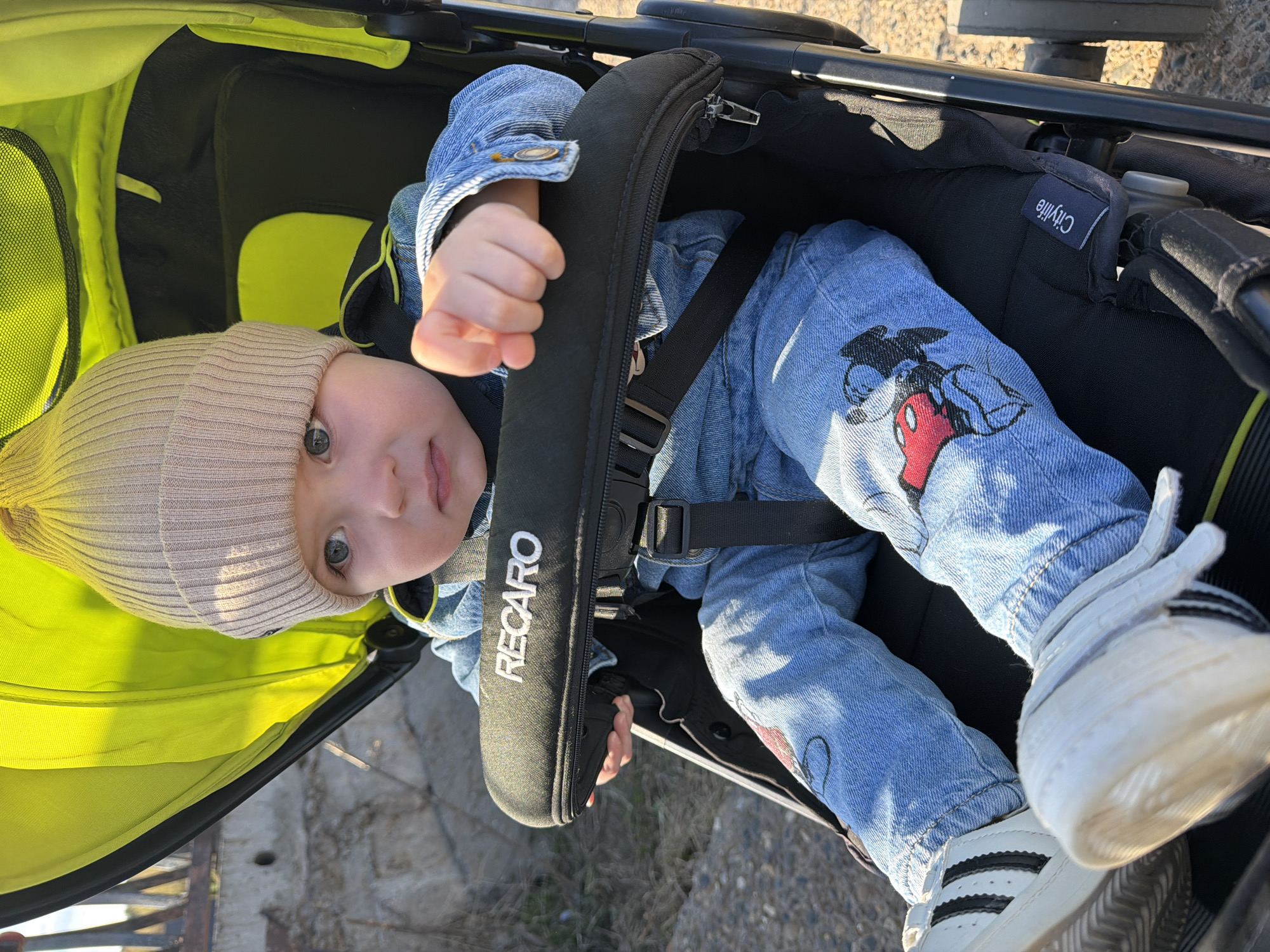 The image shows a baby sitting in a stroller. The baby is wearing a beige beanie, a denim jacket, and blue jeans with a Mickey Mouse design on the leg. The baby is secured in the stroller with a harness. The stroller has a green canopy and a black frame with the brand "RECARO" visible on the harness padding. Another part of the stroller says "Citylife." The stroller is on an outdoor surface, possibly pavement. The location in the city of Votkinsk, Russia cannot be confirmed.