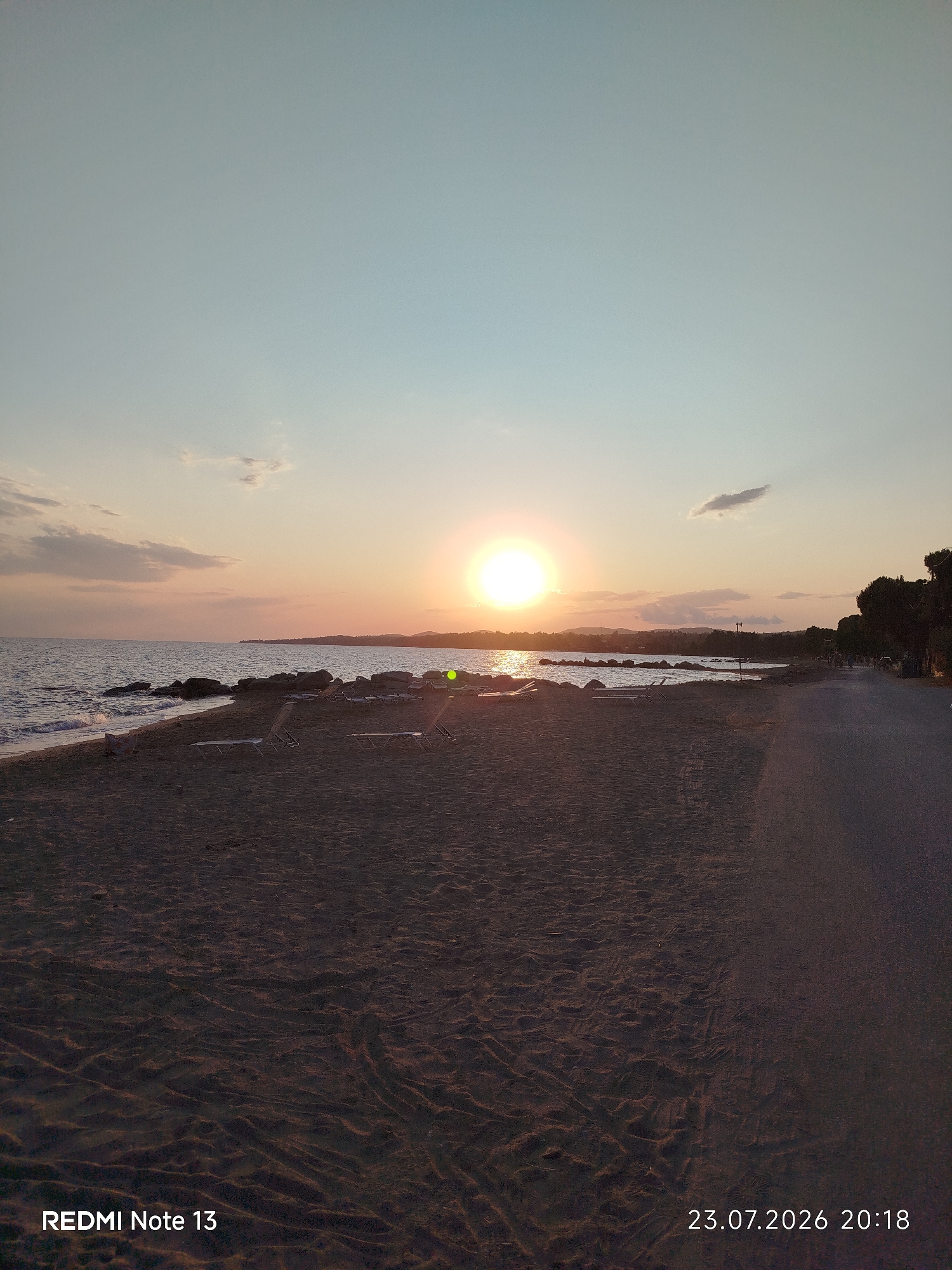 This image depicts a serene beach scene at sunset in Sithonia Municipal Unit, Greece. The sun, a bright orb, is low on the horizon, casting a warm glow across the sky and reflecting on the calm sea. The sky transitions from a pale blue at the top to soft oranges and pinks near the horizon, with a few wispy clouds adding texture.
The foreground is dominated by a sandy beach, showing footprints and the texture of damp sand near the waterline. Several empty lounge chairs are scattered across the sand, suggesting a quiet, uncrowded moment. The sea is gently lapping against a rocky shoreline, with some larger rocks visible near the water's edge.
To the right of the beach, a paved path or road runs parallel to the coast. A line of trees with dense foliage borders this path, and in the distance, there are faint silhouettes of hills and buildings. A few figures are visible walking or standing along the path, adding a subtle human presence to the tranquil landscape. The overall atmosphere is peaceful and evokes a sense of relaxation associated with the end of a day at the beach.
The visible text in the lower left corner reads "REDMI Note 13" and in the lower right corner, "23.07.2026 20:18", indicating the device used and the date and time the photo was taken.