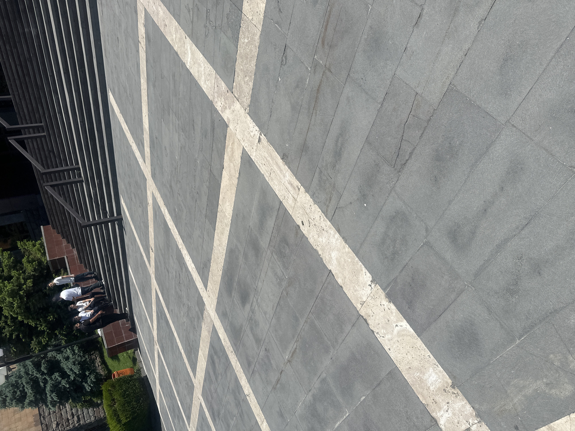 The image shows an outdoor view of a patterned stone surface adjacent to a building, potentially located in Yerevan, Armenia. The surface is composed of grey square stones arranged in a grid pattern, with off-white stripes bisecting the squares diagonally.
On the left side of the image, steps lead to a building with a linear facade. There are at least four individuals located near the base of these steps, near some bushes and trees. The people are wearing light-colored tops. It is difficult to tell what they are doing.
The scene is well-lit, suggesting daytime. The image is taken from an elevated perspective.