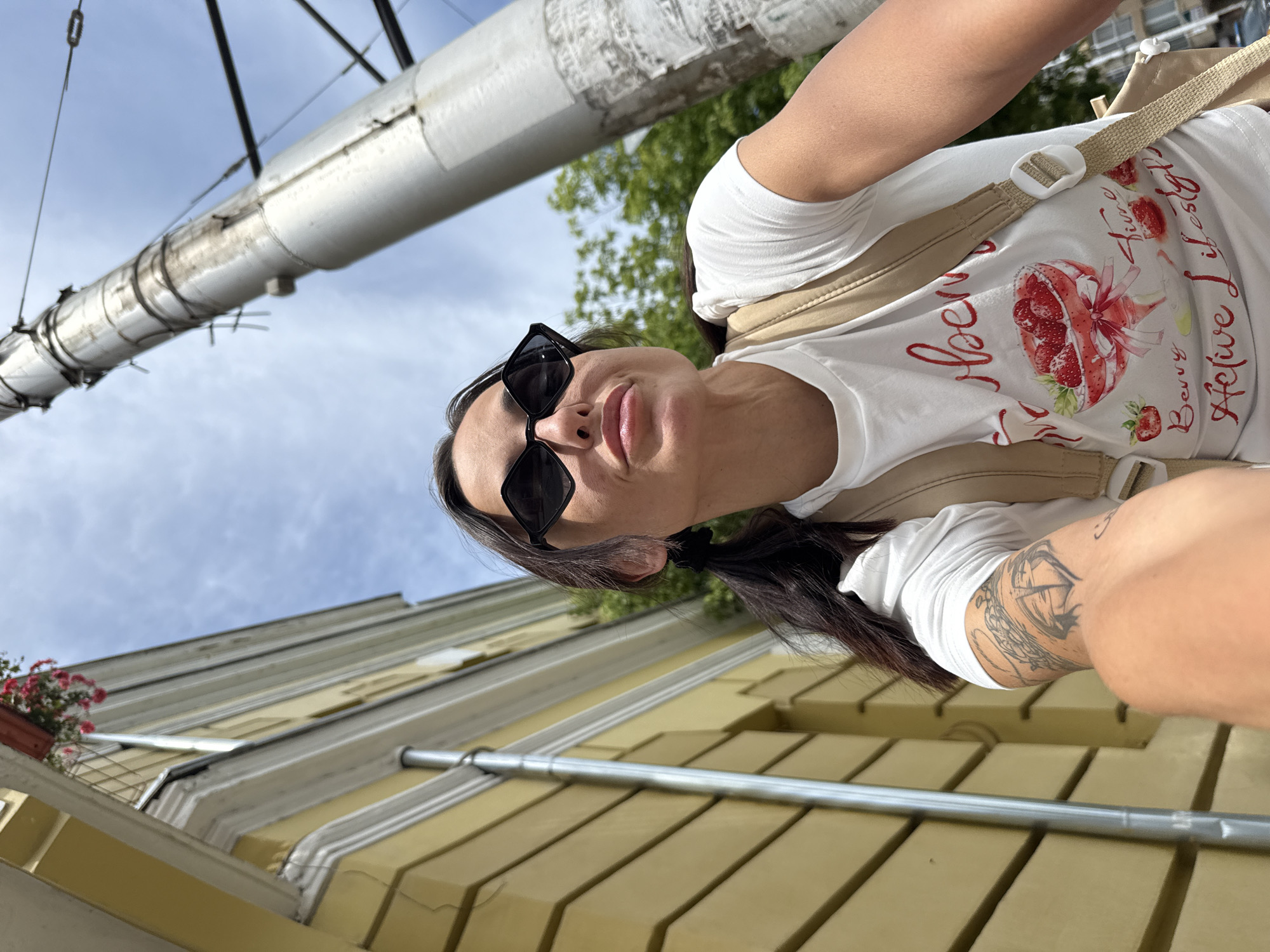 A woman is taking a selfie outdoors, possibly walking or cycling, as indicated by the angled perspective and her backpack. She has long dark hair, wears black sunglasses, and a white t-shirt with a strawberry graphic and the text "Berry Lifestyle". She has a tattoo on her arm. The background shows a light blue sky with some clouds, a large cylindrical object, and a building with yellow siding. A potted plant with red flowers is visible on a ledge. The scene appears to be set during the daytime with fair weather. The image is oriented sideways.