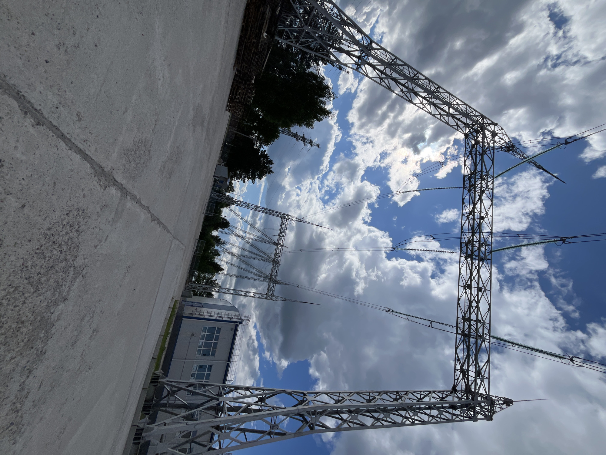 The image shows an industrial area with power transmission towers against a partly cloudy sky. In the foreground, there is a concrete ground with a visible seam. To the left, a stack of wooden pallets is present, partially obscured by trees. In the mid-ground and background, several large metal power line towers are visible, supporting electrical wires. A modern industrial building with a silver facade and blue trim, featuring several windows, is situated behind some of the towers. The scene suggests an electrical substation or a power generation facility located in or near the city of Chaikovsky, Russia. There are no people visible in the image.