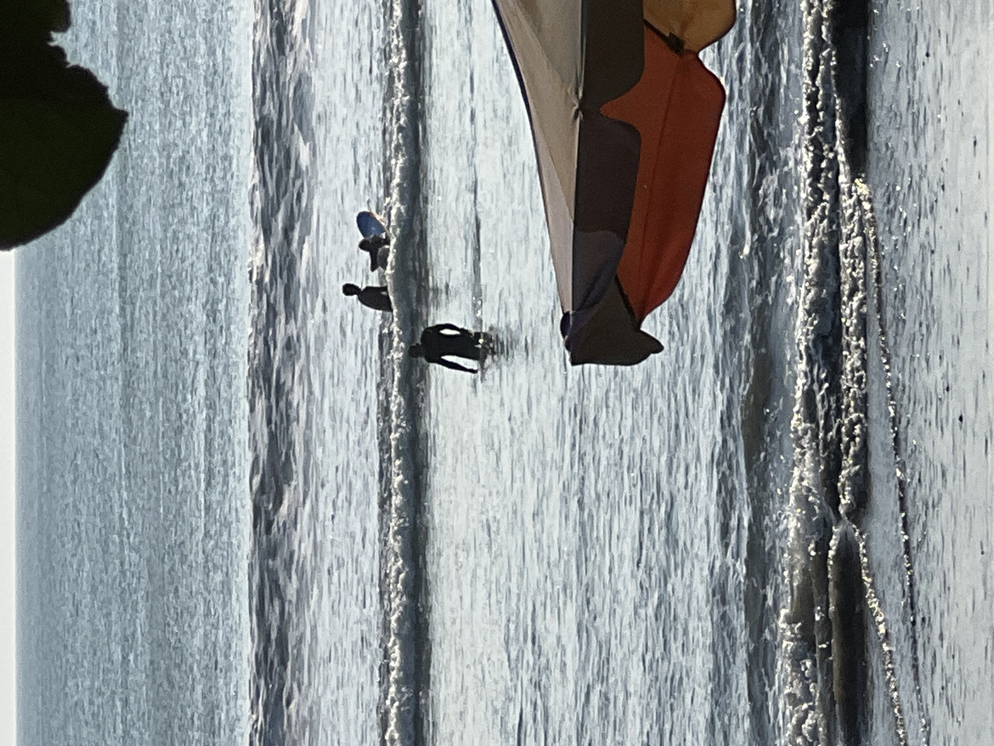 In Kuta, Indonesia, this image captures a serene beach scene with surfers in the water. In the foreground, a portion of an umbrella is visible, casting a shadow over the glistening waves. Further out, a surfer in a dark wetsuit stands in the shallow water, facing away from the camera. In the mid-ground, two silhouetted figures are engaged in surfing activities, one on a blue surfboard. The ocean stretches out to the horizon, with gentle waves rolling in, reflecting the soft, natural light. The atmosphere is tranquil and evokes a sense of a peaceful day at the beach.