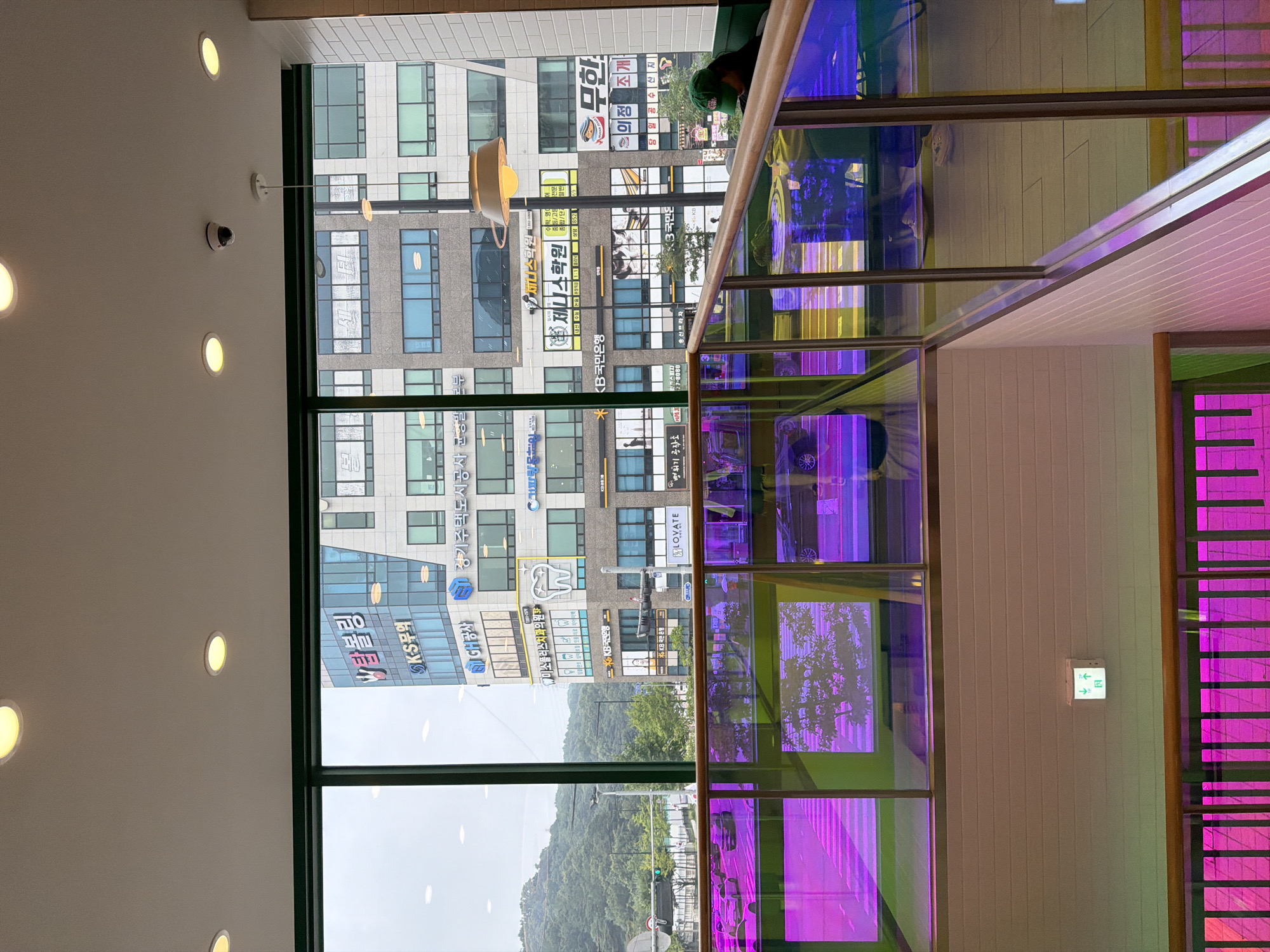 The image shows an interior view from a modern building in Uijeongbu-si, South Korea. The perspective is from an elevated position looking out through large windows. Outside, several multi-story buildings are visible with various signs in Korean. A lush green hillside forms the backdrop.
The interior features a staircase or multi-level area with reflective glass railings that display vibrant pink and purple light. The glass is also tinted with blue and yellow hues. The walls are lined with white textured tiles. In the foreground, there are glimpses of people seated in the lower section of the building, some wearing casual clothing. One person is wearing a green baseball cap and a dark jacket.
Above the windows, a security camera is mounted on the ceiling, along with several recessed circular lights. The overall atmosphere suggests a commercial or public space, possibly a cafe, restaurant, or shopping area, with a contemporary and colorful design.
