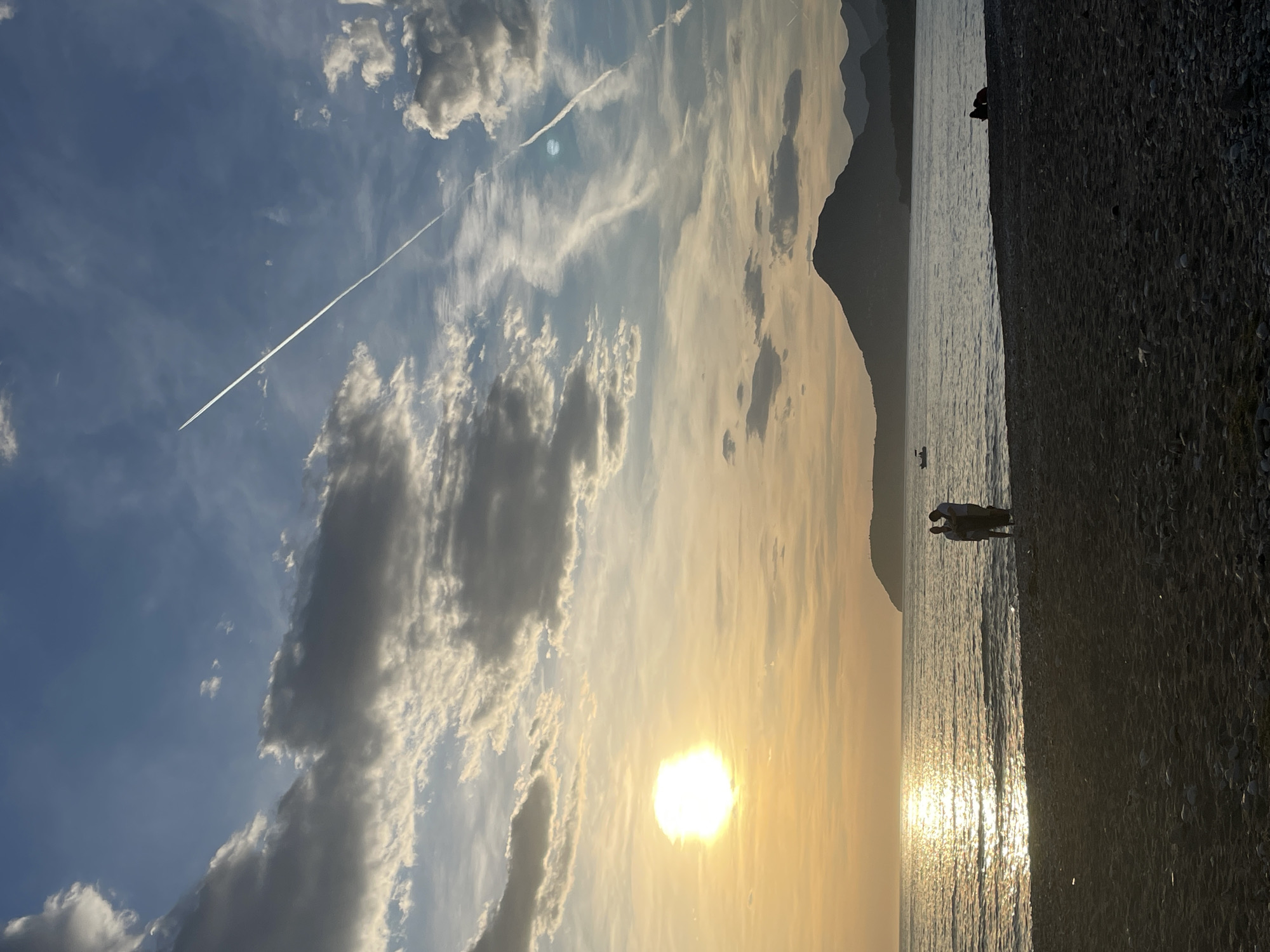 The image depicts a sunset on a beach near Bar, Montenegro. In the foreground, there's a dark pebble beach transitioning into a sea reflecting the golden sunlight. Two individuals are standing by the water's edge. Further out, a small boat is visible. In the background, there's a silhouette of a mountain range. The sky is filled with clouds, and a contrail is visible from an aircraft. The sun is setting, casting a warm light across the scene.