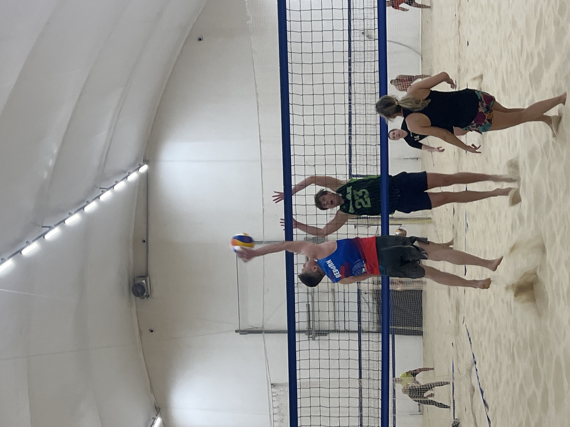 A beach volleyball game is in progress indoors, taking place on a sand-covered court. The net is stretched across the center, dividing the two teams. On one side of the net, a male player wearing a blue and red jersey with the name "REBORN" on it, is in the process of hitting the volleyball with his right hand. His opponent, a male player wearing a green jersey with the number "23", is positioned at the net, attempting to block the hit. In the background, a female player wearing a black tank top and colorful shorts is also on the same side as the green jersey player. Other players are scattered on the court. The environment is a bright indoor arena with a white, dome-shaped ceiling. Based on the jersey name and assuming the photo location is St. Petersburg, this scene is possibly located inside one of the beach volleyball facilities.