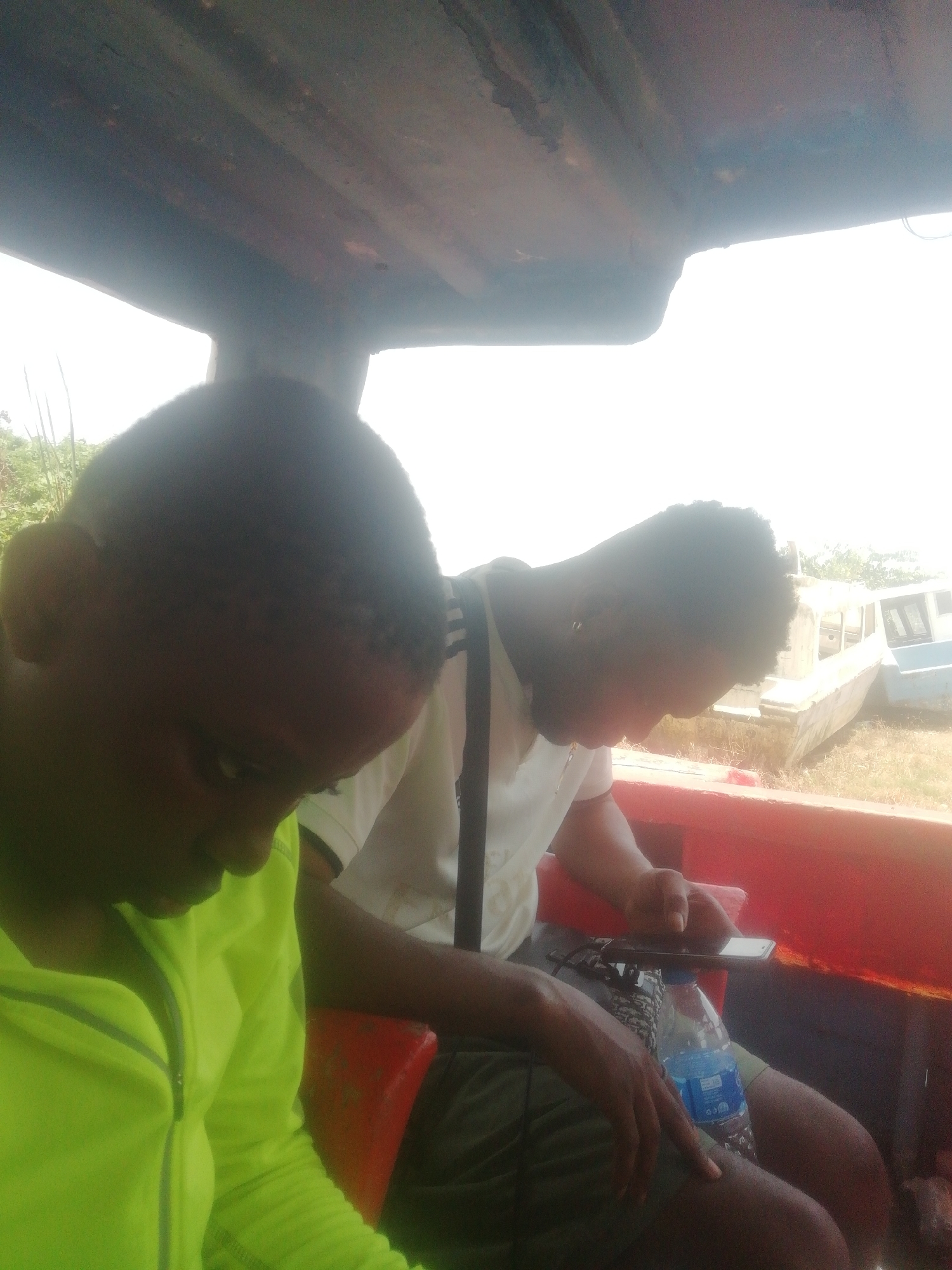 Two people are seated inside a boat. One person is wearing a bright yellow jacket, while the other is wearing a white shirt with dark straps, looking at a mobile phone placed over a water bottle. The background shows old boats resting on land with vegetation. Location: Unknown, Unknown.