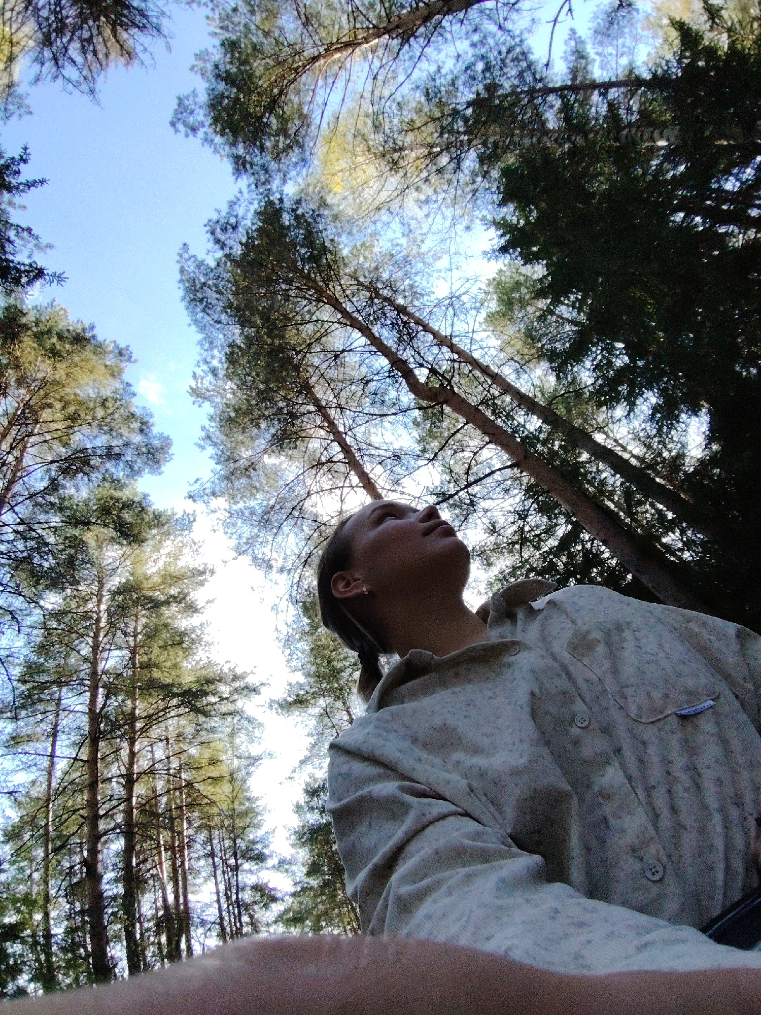 The image shows a low-angle shot of a person looking up at tall pine trees in what appears to be a forest. The sky is visible through the canopy, and it is partially clear with some clouds. The person is wearing a light-colored, buttoned shirt. The scene gives the impression of being in a natural environment, specifically a forest near the city of Марковский, Russia.