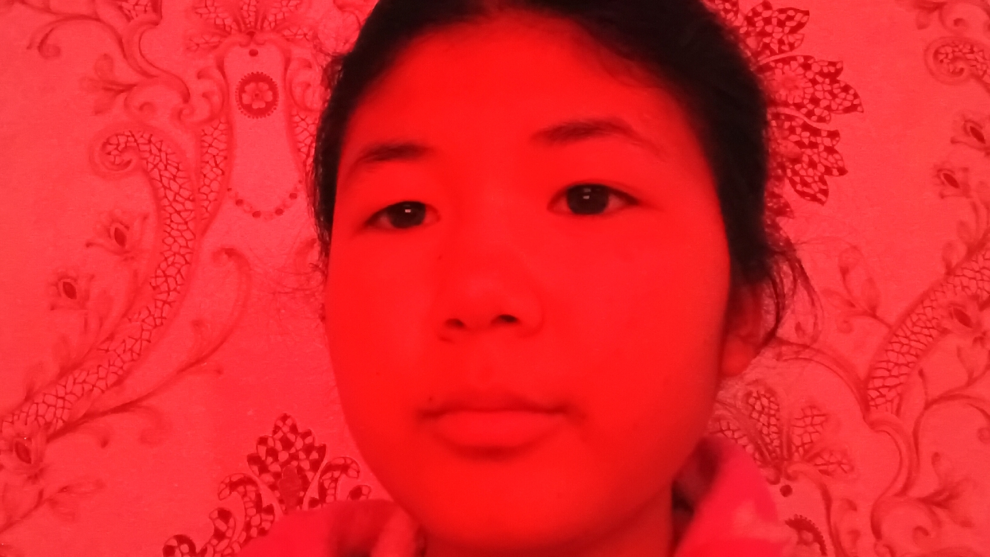 The image shows a close-up, headshot of a young East Asian female, likely a teenager, illuminated by a strong red light source, giving the entire scene a crimson hue. She is positioned in the foreground, with a red-patterned wallpaper serving as the background. The wallpaper features an ornate, possibly floral or damask-like design, with repeating motifs and swirling lines. The lighting obscures many of the finer details of her facial features, but her dark eyes, eyebrows, nose, and lips are discernible. Her hair, dark and appearing to be straight, frames her face. The image appears to be a selfie, as it is framed from her perspective. There is no visible indication of the specific location within Uchkun, Kyrgyzstan, beyond the generic wallpaper pattern.