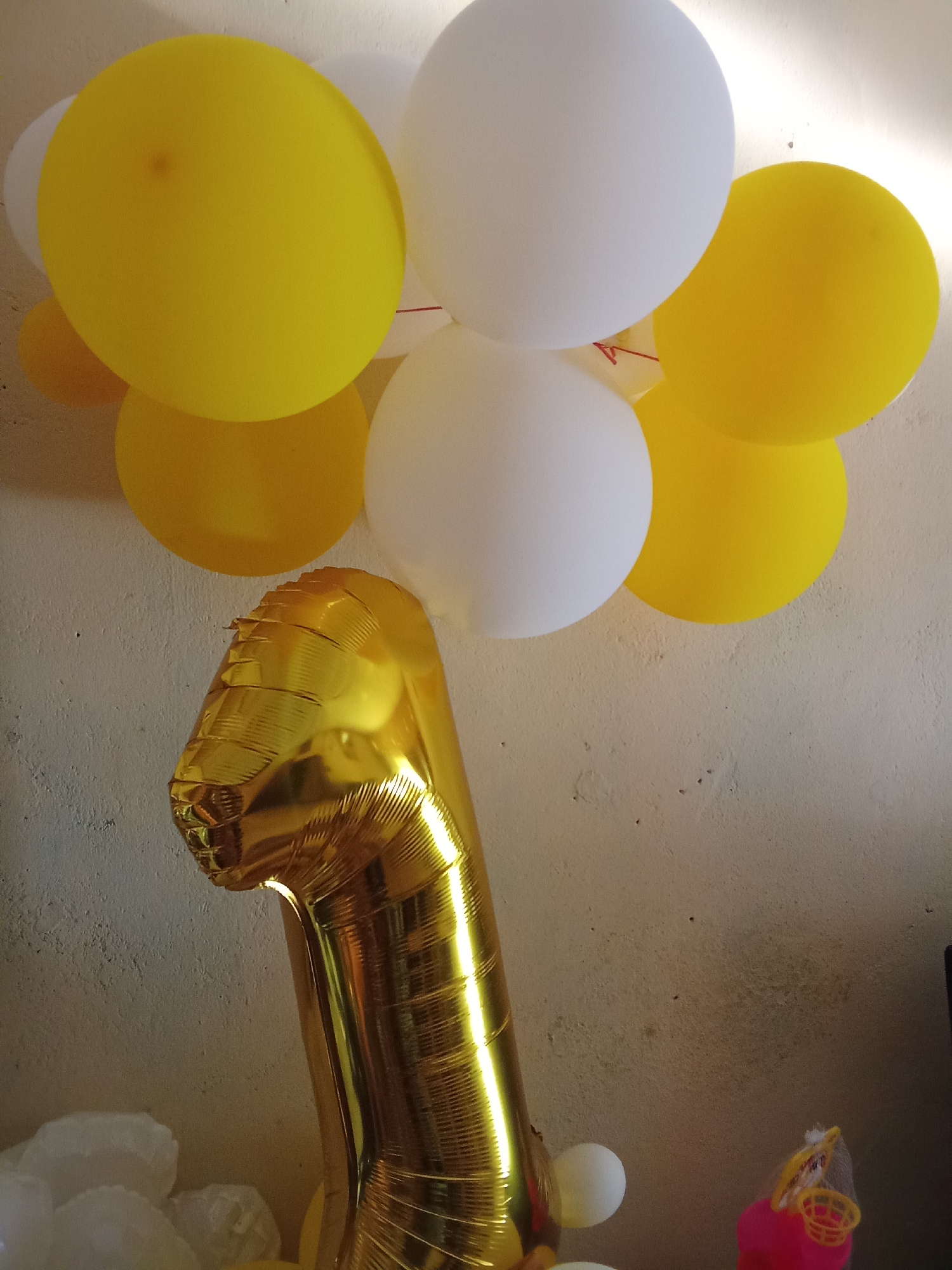 A close-up, low-angle shot shows a collection of yellow and white balloons floating against a textured, light-colored wall. Prominently featured in the foreground is a large, golden Mylar balloon shaped like the number "1." The balloons are clustered together, with several yellow and white ones filling the upper portion of the frame. The wall appears to be concrete or plaster, showing some imperfections. In the bottom right corner, a small, pink toy with a yellow basketball hoop and net is visible, partially obscured. The lighting suggests an indoor environment, possibly a home or event space. The scene does not provide any specific indicators of its location within Tanah Mas, Indonesia.