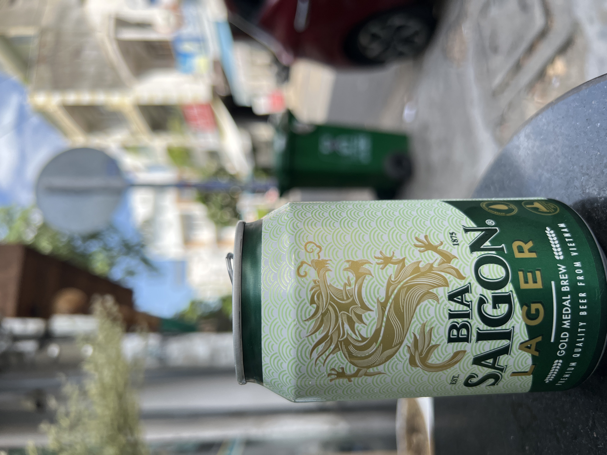 A Saigon Lager beer can is prominently featured in the foreground of the image, placed on a dark, textured surface that appears to be a table. The can has a light green background with a repeating pattern of light green waves, and a large golden dragon design. Text on the can includes "BIA SAIGON", "LAGER", "EST. 1875", "GOLD MEDAL BREW", and "PREMIUM QUALITY BEER FROM VIETNAM". The can's lid is visible, as is the pull tab.
The background is out of focus but suggests an outdoor urban setting. Buildings with multiple stories are visible, with balconies and windows. There are hints of a street with some vegetation and a red vehicle to the right of the can. A green trash bin is also present in the background. The lighting indicates it is daytime, likely sunny. The overall impression is a casual, outdoor beverage setting in a Vietnamese city.