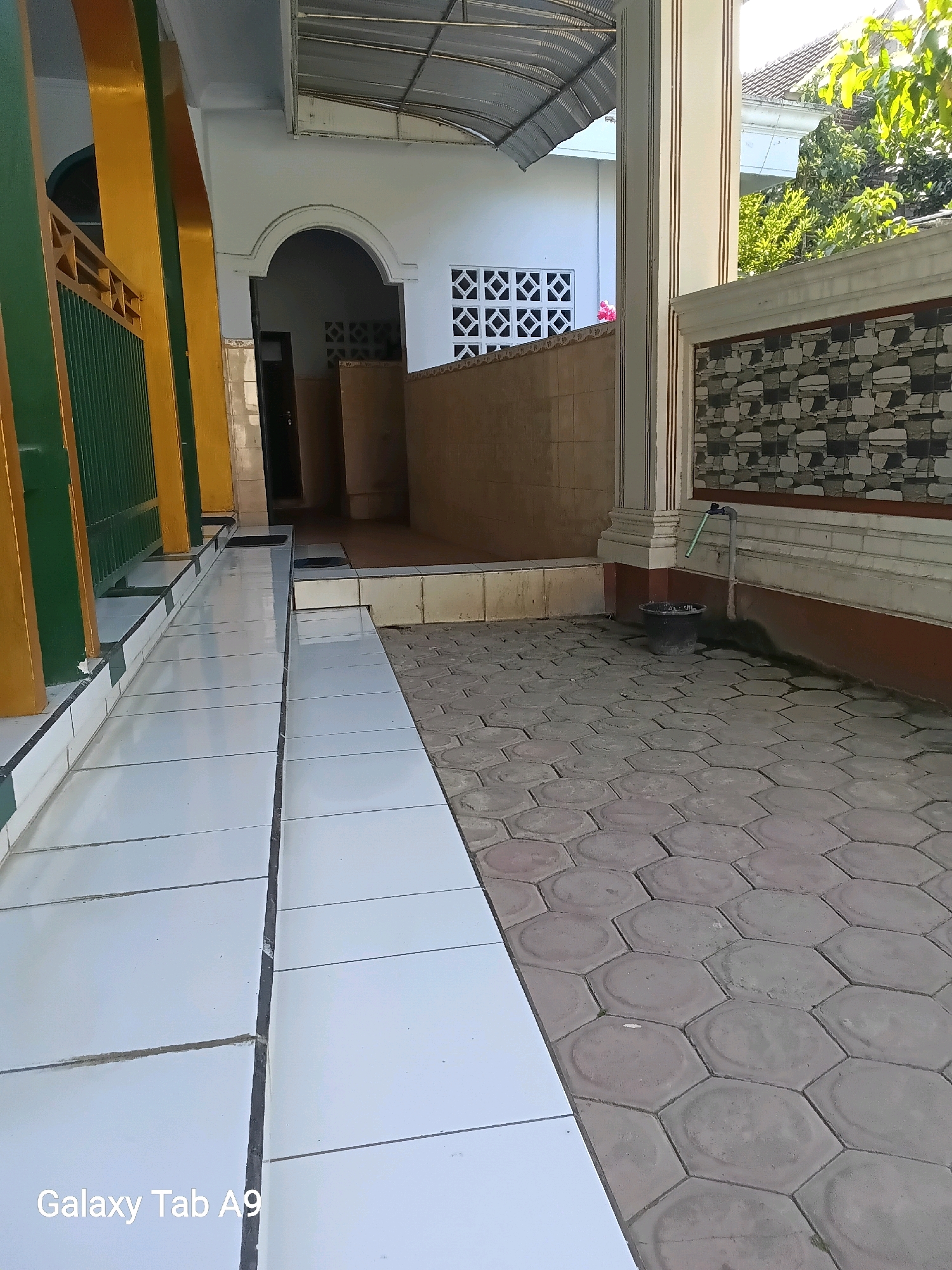 An outdoor walkway and patio area are shown. To the left, a ramp with white tiles and dark grout leads towards a building entrance with an arched doorway. Yellow and green pillars frame the walkway. To the right, hexagonal concrete pavers form the patio. A faucet with a green pipe is visible on a wall with decorative stone and striped detailing. A black bucket sits on the patio beneath the faucet. The scene is bathed in sunlight, casting shadows on the ground. The background includes a building with a metal roof and trees. The bottom left corner of the image displays "Galaxy Tab A9".