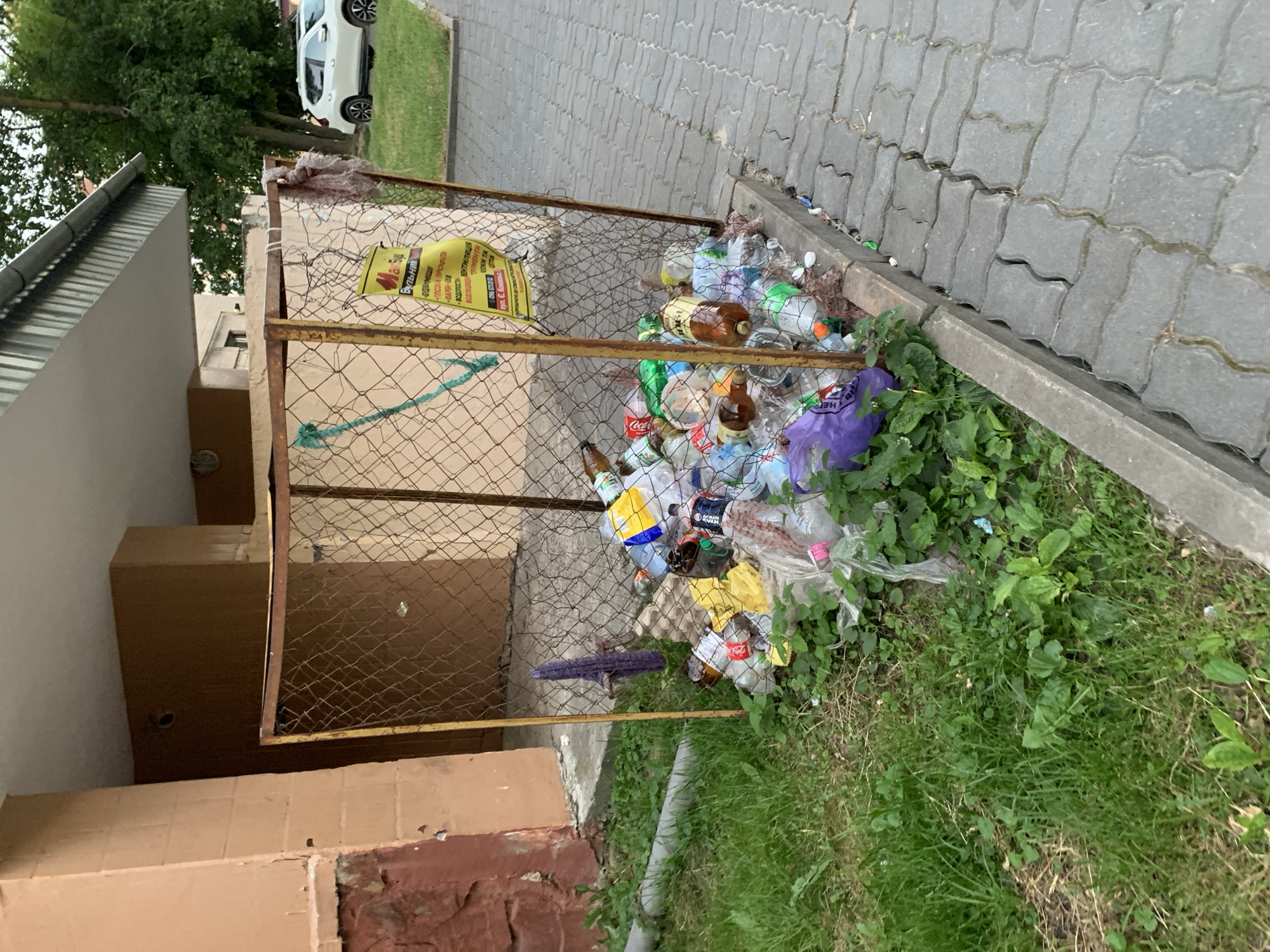 This media file shows an outdoor scene in Ternopil, Ukraine, likely on a cloudy day. The primary subject is a metal cage dumpster filled with plastic bottles and other recyclable waste. The cage is old and rusted, with a yellow sign attached to it. The sign contains text in Cyrillic, with some legible words such as "Coca" and "Бульний" (Bulʹnyy). The dumpster is situated next to a grey, patterned paved walkway and a patch of green grass with weeds. In the background, a white car is partially visible, along with trees and a building. The overall impression is one of neglect and uncollected waste.