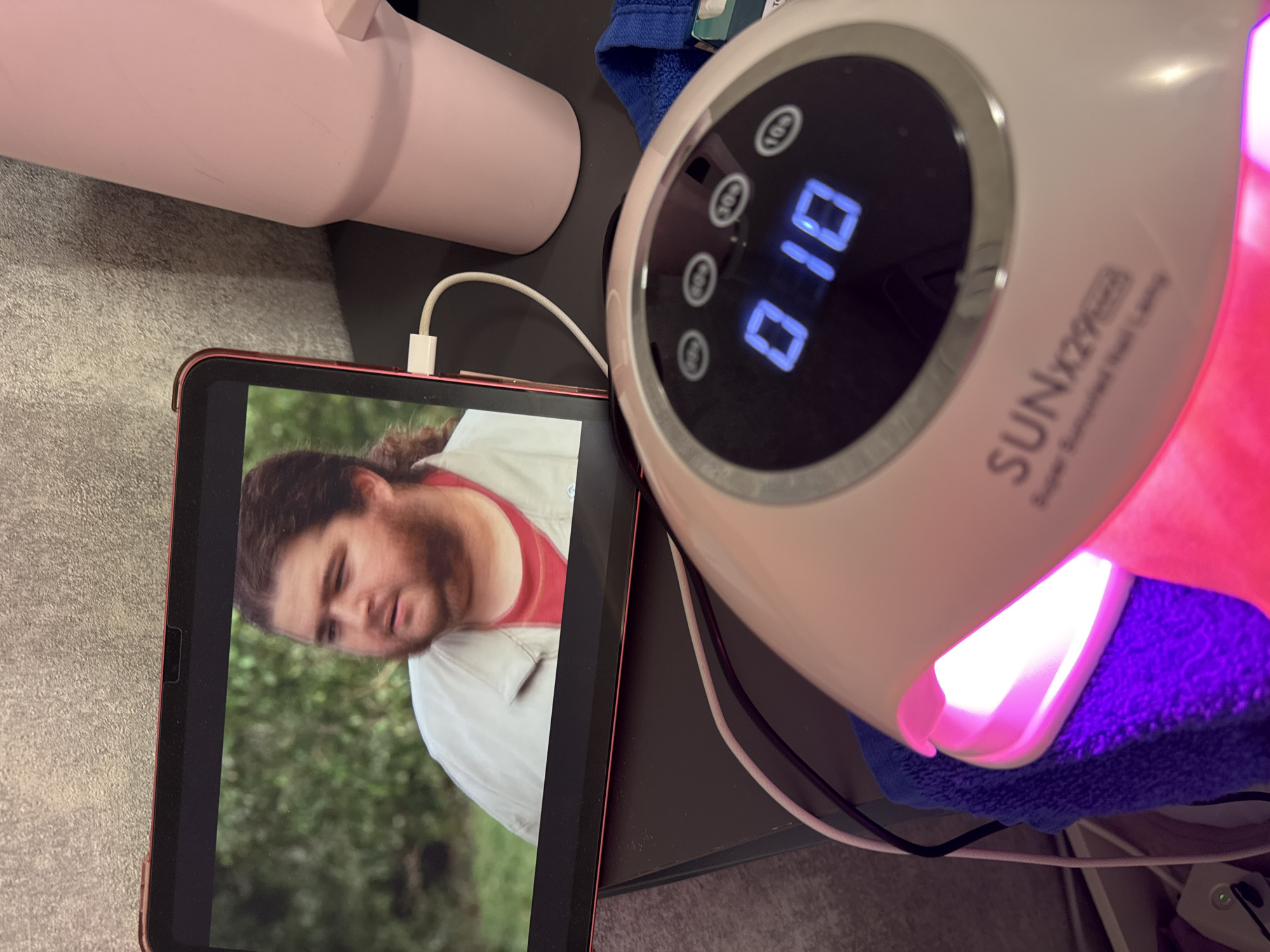 A high-angle shot shows an indoor scene with several objects arranged on a gray surface, likely a table or desk.
A pink tumbler sits in the upper-left. A red-cased tablet displays a man with brown hair, a beard, and a red shirt under a light-colored jacket; foliage is visible in the background, suggesting an outdoor setting. A charging cable is plugged into the tablet.
To the right of the tablet is a white SUNx29 nail lamp, illuminated with a pinkish-purple light shining on a blue towel. The lamp's display shows the number "010".
The scene's location within Plovdiv, Bulgaria, cannot be determined from the provided image.