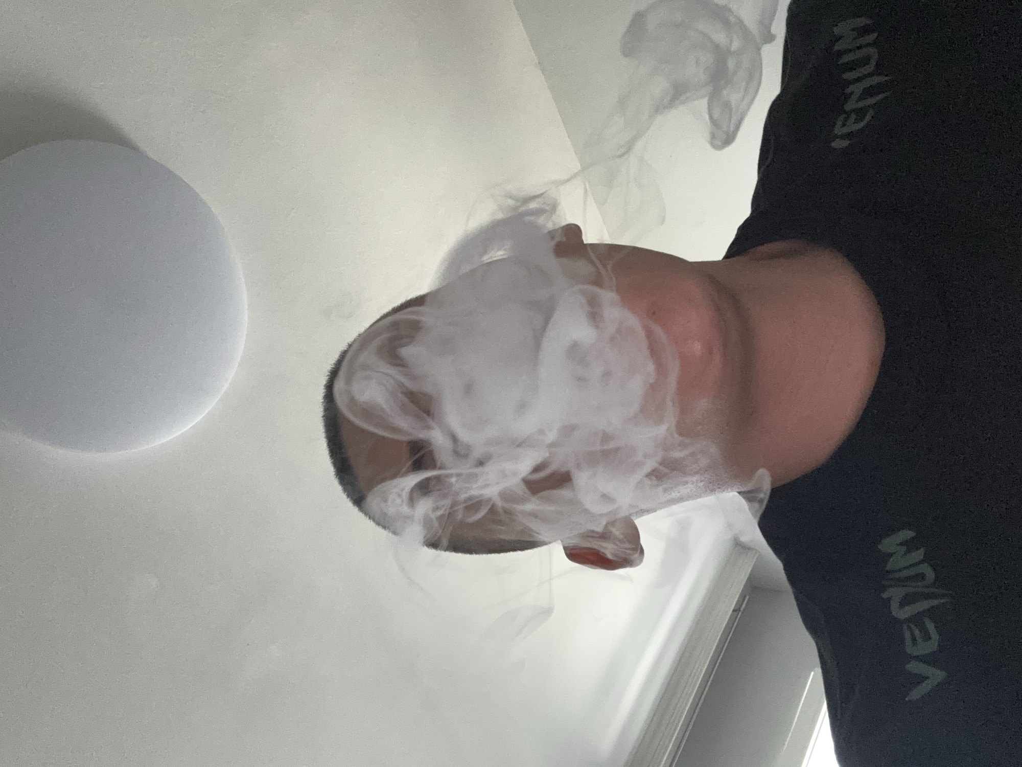 The image, taken in Plovdiv, Bulgaria, is a low-angle, close-up shot of a person indoors, rotated 90 degrees counter-clockwise. The primary subject is a person, likely male, whose head (top and side), part of the face (chin and jawline), and a portion of the upper body/shoulder are visible. The person has short, dark hair and is wearing a black garment, possibly a t-shirt or hoodie, with the brand name "VENUM" visible in grey letters along the side.
The central activity depicted is the person exhaling a large, dense cloud of white vapor or smoke, which envelops and partially obscures their face and the area immediately around their head.
In the background, to the upper left (when oriented correctly), a round, flat, white or light grey ceiling light fixture is visible. The rest of the background consists of a plain, light-colored ceiling. The setting appears to be a modern indoor space. No specific time of day or emotional state is discernible from the limited view.