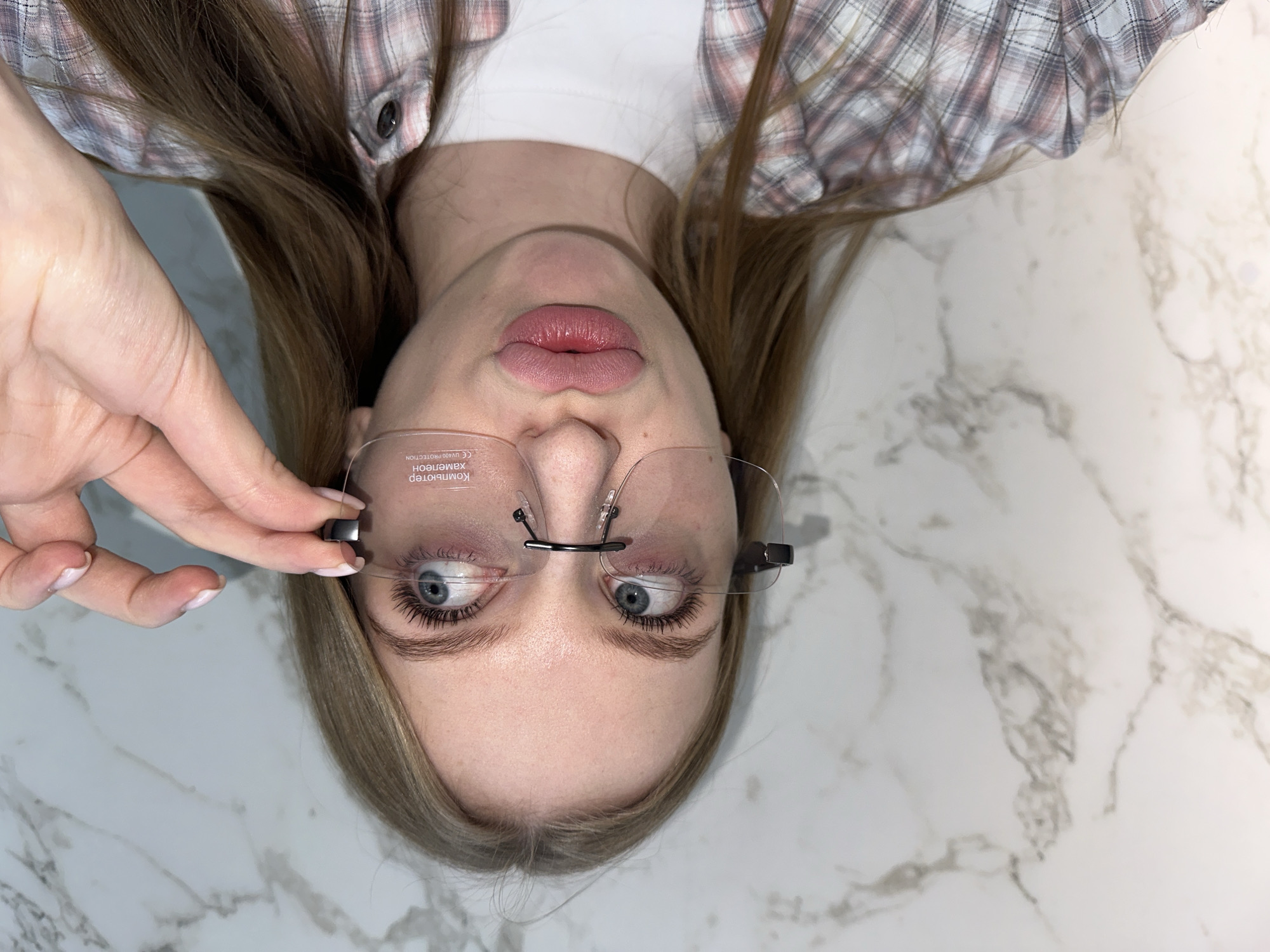 A young adult female is depicted in a close-up, chest-up indoor shot, positioned against a white marble-patterned wall. The specific location within the city of Chaikovsky, Russia, is not discernible from the image. She has fair skin, light brown hair, and blue eyes, with visible mascara on her eyelashes. Her lips are pursed forward in a "duck face" expression. She wears a light-colored plaid shirt, predominantly pink and grey, over a white t-shirt. She is wearing rimless eyeglasses with clear lenses. The right lens (from the viewer's perspective) displays faint white text in Cyrillic, "Компьютер хамелеон" (Computer Chameleon), and "CE UV400 PROTECTION". Her left hand, with light pink manicured fingernails, is holding the black temple piece of the eyeglasses on the right side of her face, near her temple. Her gaze is directed upwards and to her right.
