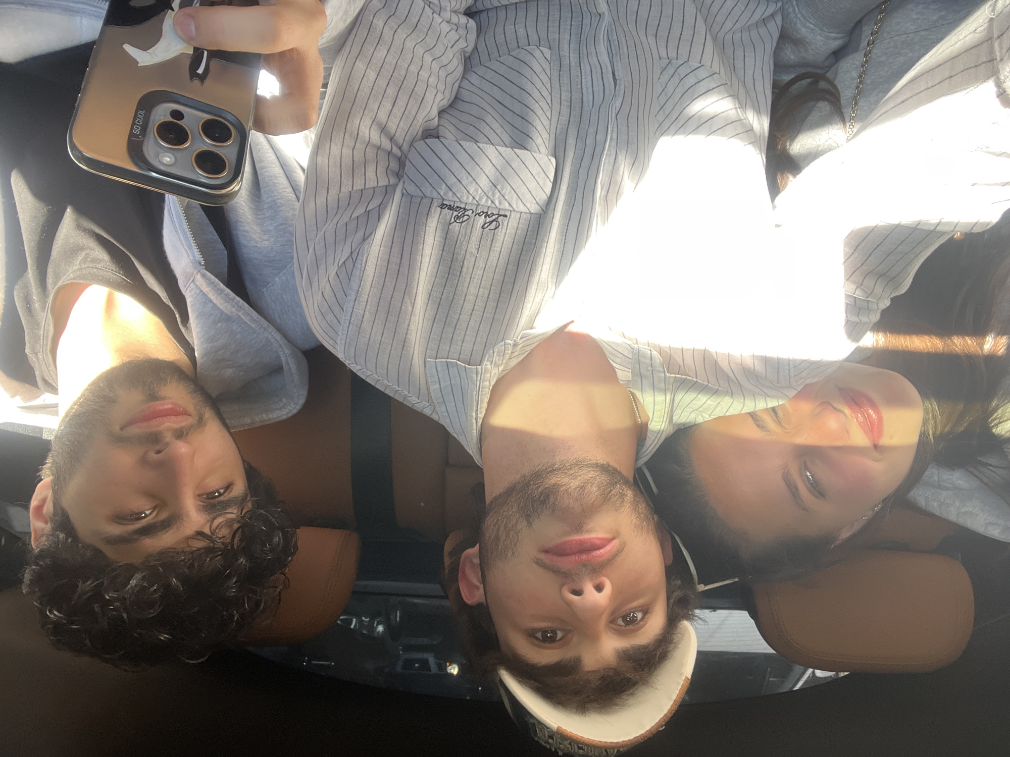 A photo shows three people in what appears to be the back seat of a car. One person holds a phone with a clear case with a graphic on it. A man on the left has dark, curly hair and wears a gray hooded sweatshirt over a black shirt. The man in the center has a beard and wears a striped shirt with the words "Lovers Era" on the pocket. A woman sits to his right and is wearing a gray hoodie. The interior of the car has brown leather seats. The background of the scene is undiscernible. The location of Yerevan, Armenia cannot be verified based on the image.