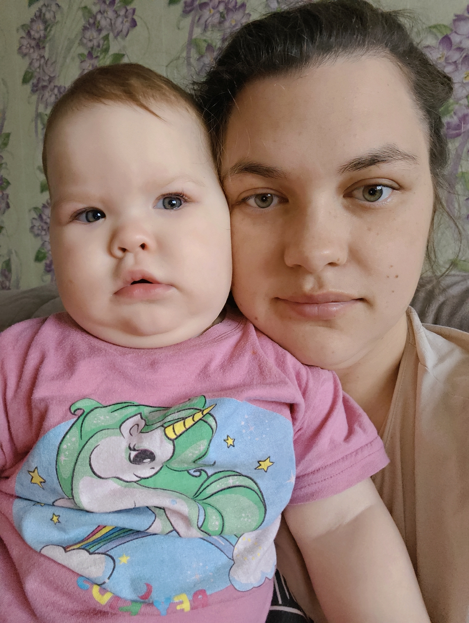 A close-up indoor shot depicts a young child and an adult female. The child, wearing a pink shirt with a unicorn graphic, is positioned on the left. The adult woman is on the right, her face close to the child's. The background is a floral patterned wallpaper. There is no indication this photo was taken in Уральское, Russia.