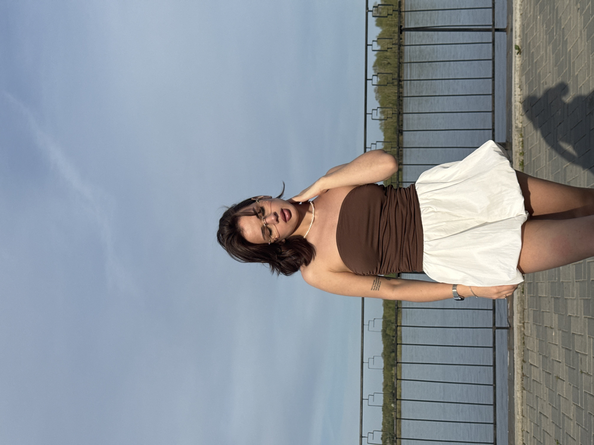 A young woman poses in an outdoor setting. She is wearing a brown strapless top and a white skirt. She has brown hair and is wearing glasses and a pearl necklace. Her right hand is touching her neck. A wristwatch is visible on her left wrist, along with a tattoo on her left forearm. Behind the woman is a body of water and a black metal railing, along with trees and foliage in the background. The setting appears to be a paved walkway or promenade. The sky is light blue. The location is Sarapul, Russia.