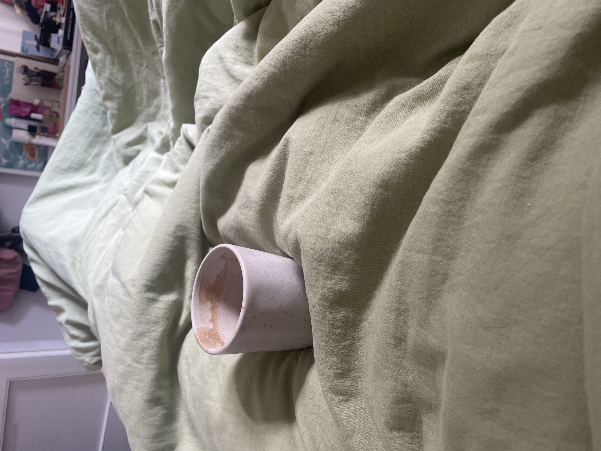The image displays an indoor scene, specifically a rumpled bed covered with light green linen bedding, located in the city of Piacenza, Italy. A light pink, speckled ceramic cup, tapered at the base and wider at the rim, is centrally positioned on the linen. The interior of the cup contains dried, light brown residue, indicative of a recently consumed beverage. The linen sheets exhibit numerous folds and creases, suggesting recent occupancy or activity. In the blurred background, to the upper left, a white door frame abuts a light-colored wall. To the upper right, various indistinct objects including bottles, what appears to be a brush holder, and other items are visible on a surface, likely a dresser or shelf. A soft, indirect light illuminates the scene, creating subtle shadows within the folds of the fabric and around the base of the cup. No individuals are present.