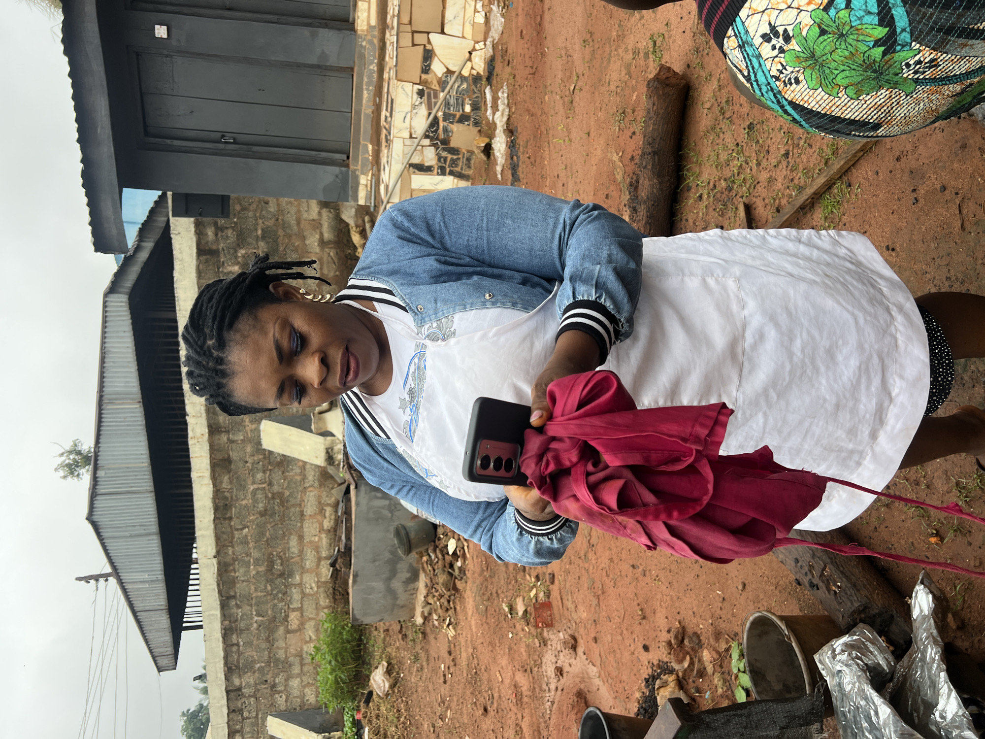 In Nkpor, Nigeria, a woman stands outdoors, wearing a white apron over a white top and a denim jacket. She holds a smartphone in her left hand and a crumpled red cloth in her right. The background features a brick wall, a grey corrugated metal roof structure, and a small dark building with a wooden door. The ground is covered in red dirt, with scattered debris, a log, and a bucket visible. The sky is overcast. Another person, partially visible from the back and wearing a colorful patterned garment, stands to the right.