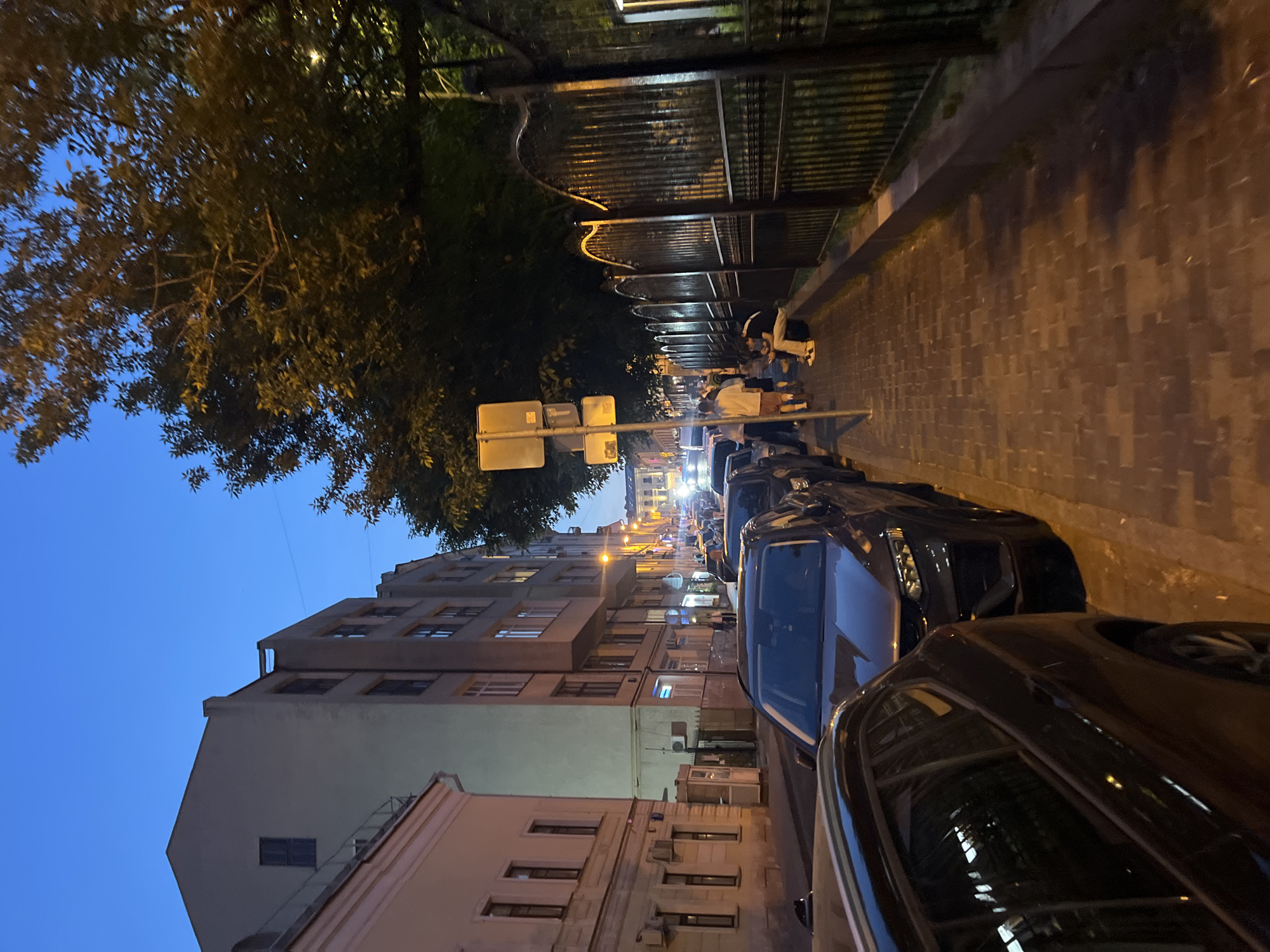 The image depicts a city street at dusk. On the left, a light-colored building with multiple windows and an arched entrance is visible. Adjacent to it is a taller, grey building with rows of windows. The street is lined with parked cars, including a dark SUV in the foreground. A sidewalk runs along the right side of the street, paved with interlocking bricks. Several individuals are walking on the sidewalk, and one person is seated on a low wall. A black metal fence with ornate detailing borders the sidewalk on the right. Streetlights are visible in the distance, illuminating the scene. The sky is a deep blue, indicating twilight.