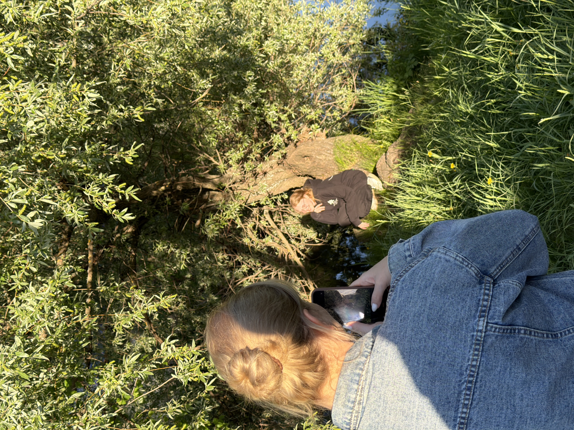The scene depicts two individuals by a water feature in Chaikovsky, Russia. One person, with blonde hair styled in a bun and wearing a blue denim jacket, is facing downwards while holding and using a smartphone. The phone's screen reflects the surrounding scenery. The second individual is lying down on a fallen tree trunk or log, clad in a dark hooded sweatshirt. Both are surrounded by lush greenery: trees and grass. The time of day appears to be sunny, casting shadows across the scene.