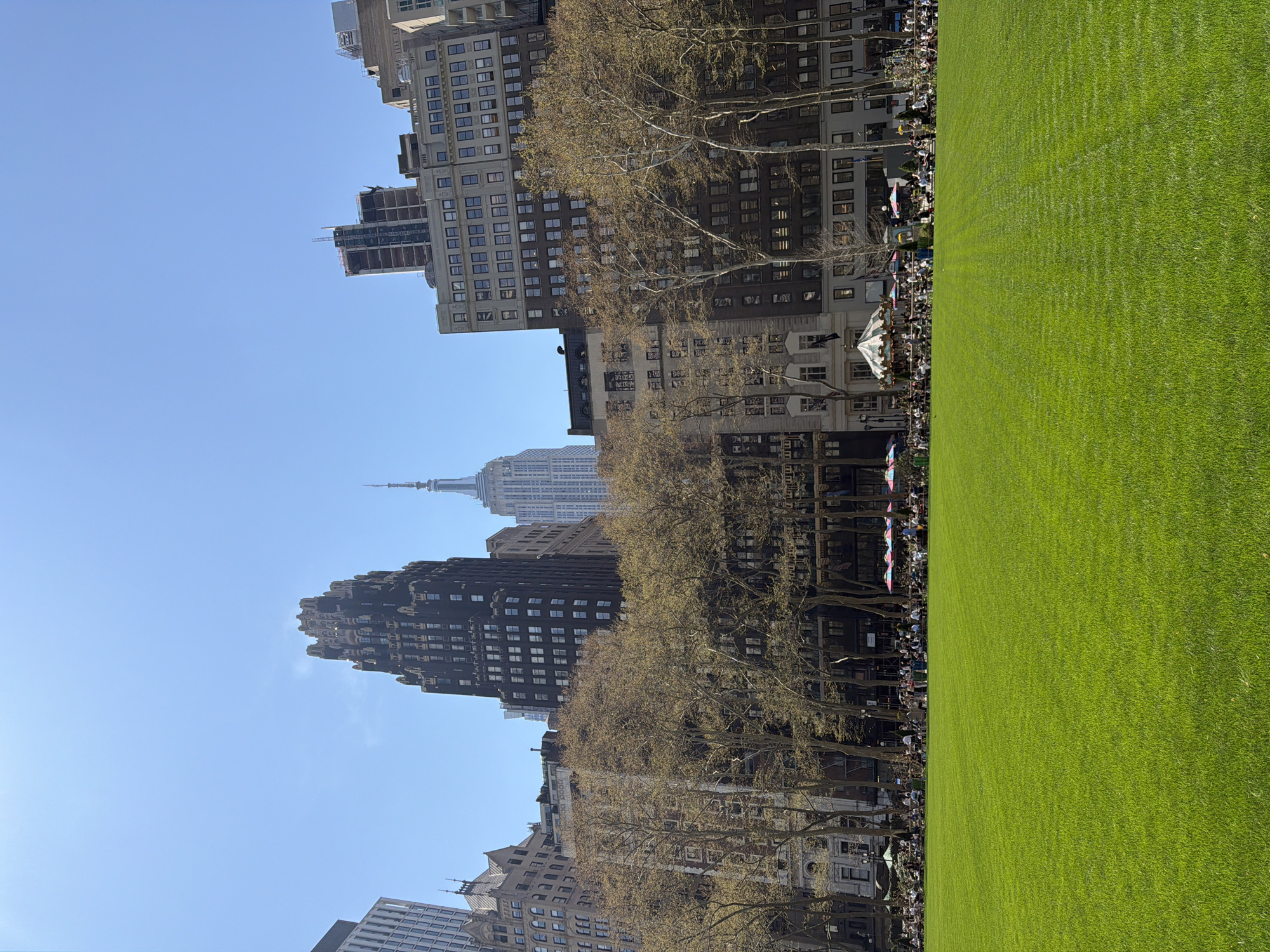 This image captures a view of Bryant Park in New York City, with the Empire State Building visible in the background. A large expanse of bright green lawn dominates the right side of the frame. Along the edge of the lawn, closest to the buildings, numerous people are seated, suggesting an outdoor event or gathering. Trees line the perimeter of the park, their branches mostly bare. The buildings in the background vary in height and architectural style, reflecting the urban landscape of midtown Manhattan. The sky is clear and blue, indicating a sunny day.