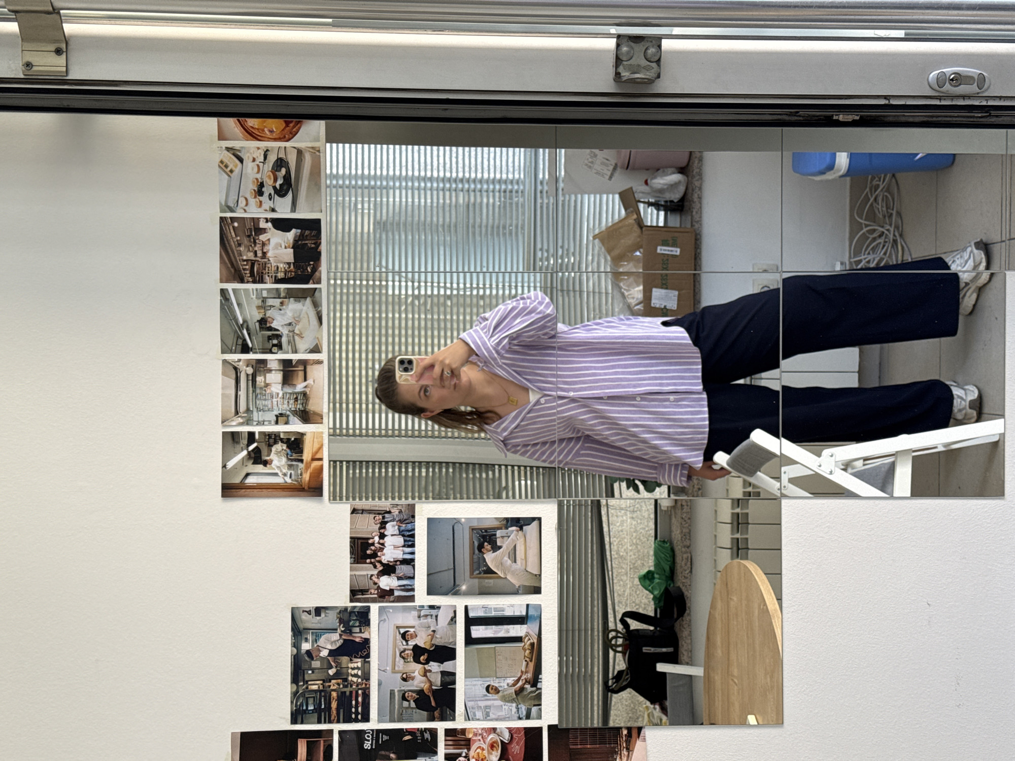 A woman is taking a selfie in a mirror. She is wearing a purple and white striped button-down shirt and dark blue wide-leg pants. The mirror reflects a cluttered kitchen or studio space, with various photographs pinned to a white wall behind her. The photographs appear to depict scenes from a bakery or kitchen, with people in chef's attire and close-ups of baked goods. In the foreground of the mirror, there is a round wooden table and a white folding chair. The background of the mirrored reflection shows a kitchen counter with items like bags, boxes, and cleaning supplies. The overall environment appears to be an indoor residential or commercial space.